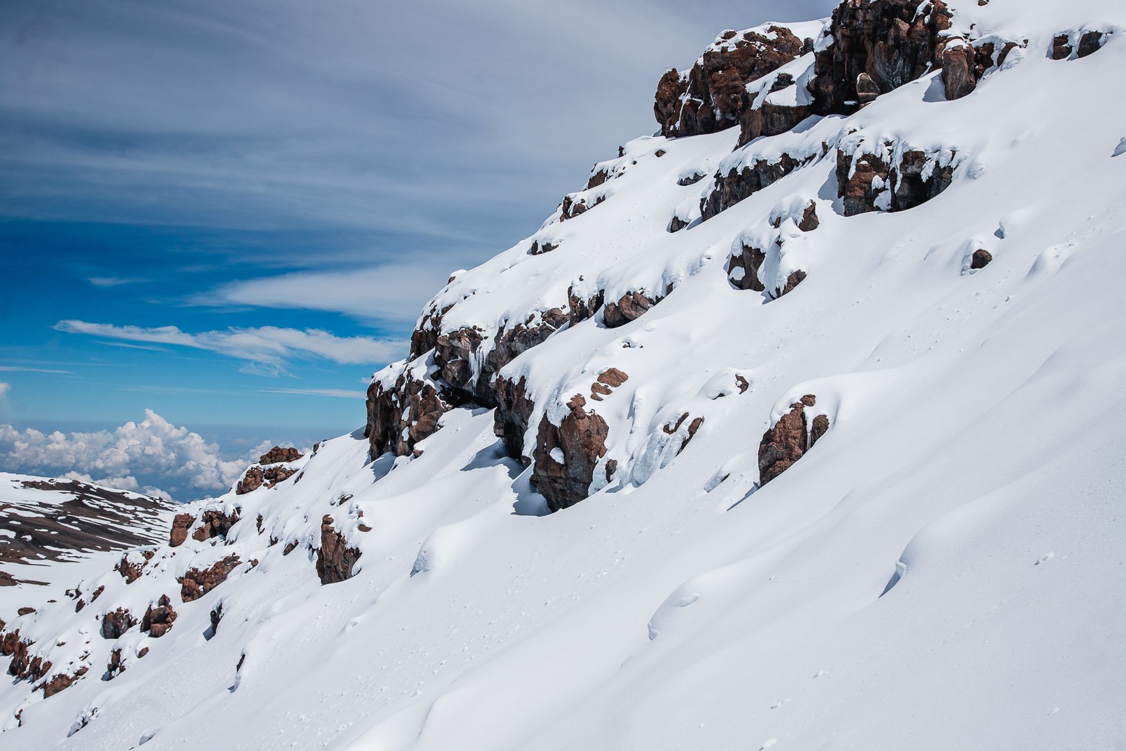 Snow-covered mountain slope with exposed rocks under a partly cloudy sky.