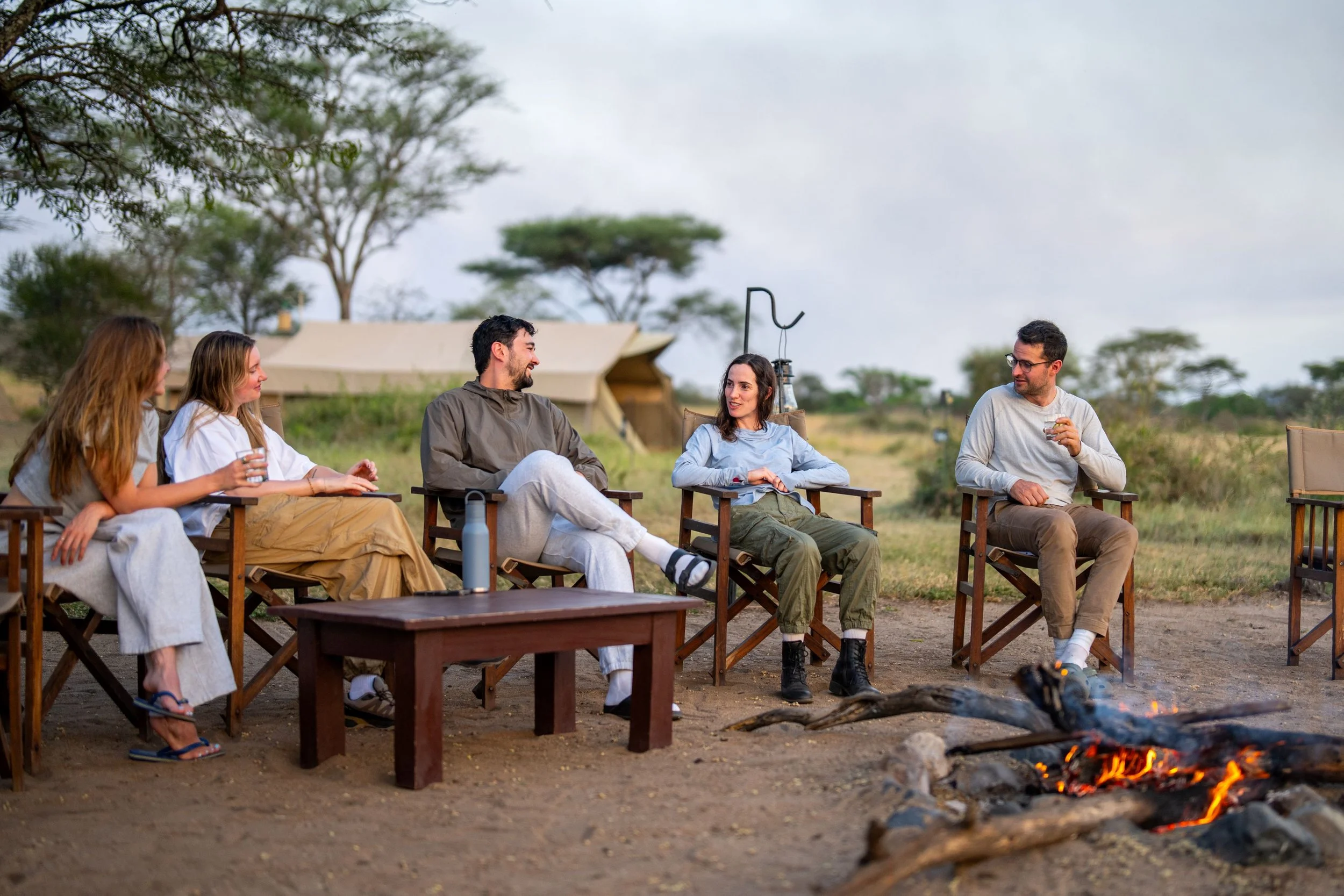 Group of five friends sitting around a campfire outdoors during daytime, with trees and tents in the background, socializing and enjoying each other's company.
