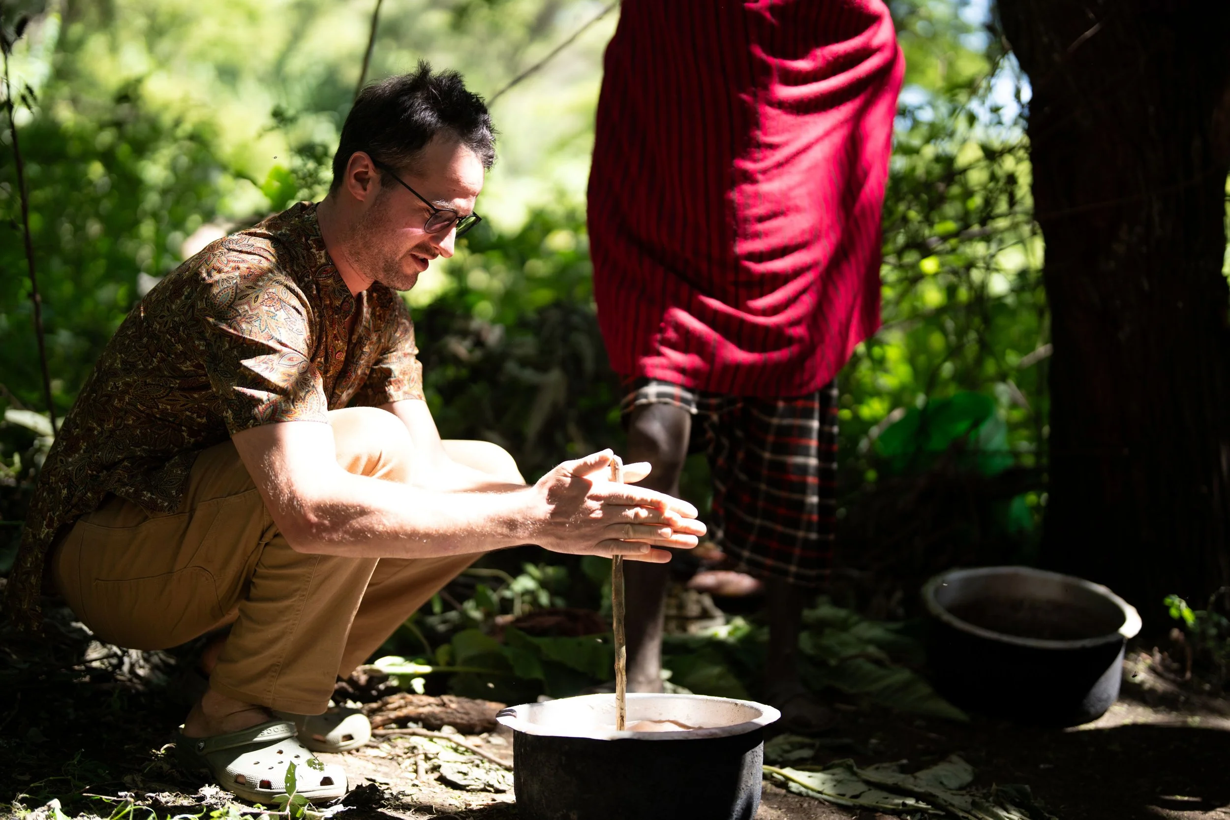 A man squatting outdoors, wearing glasses, a patterned shirt, and beige shorts, working on a small tree or plant. Another person, partially visible, stands nearby wearing a red shirt and checkered shorts. The setting is a lush green forest or garden with sunlight filtering through the trees.