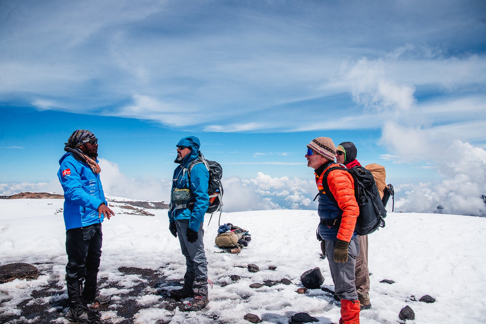 Four climbers standing on snow-covered landscape with backpacks, wearing winter gear, and talking to each other under a blue sky with clouds.