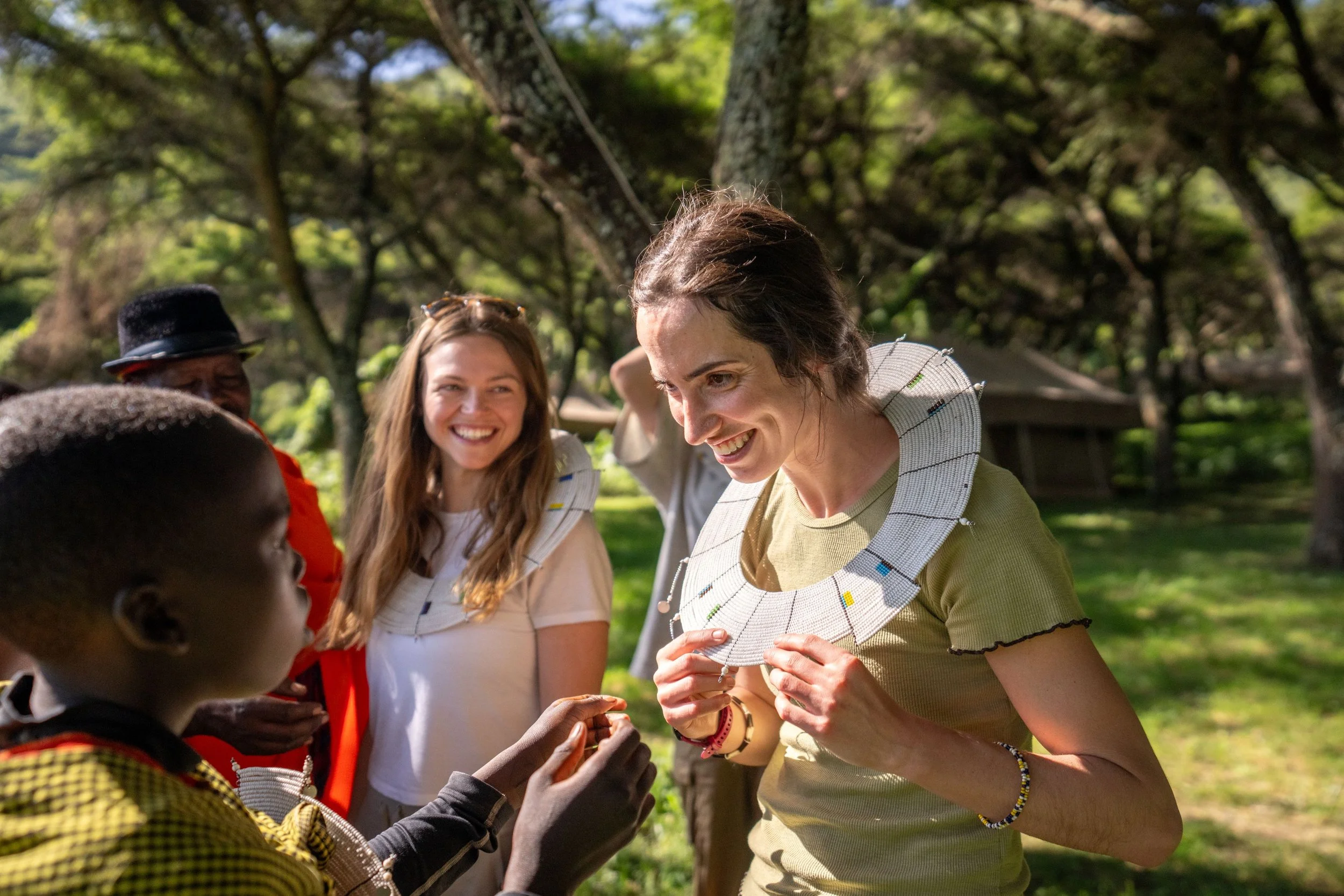 Group of people outdoors, engaging in a cultural or ceremonial activity, with a woman holding a beaded necklace and others smiling and participating.