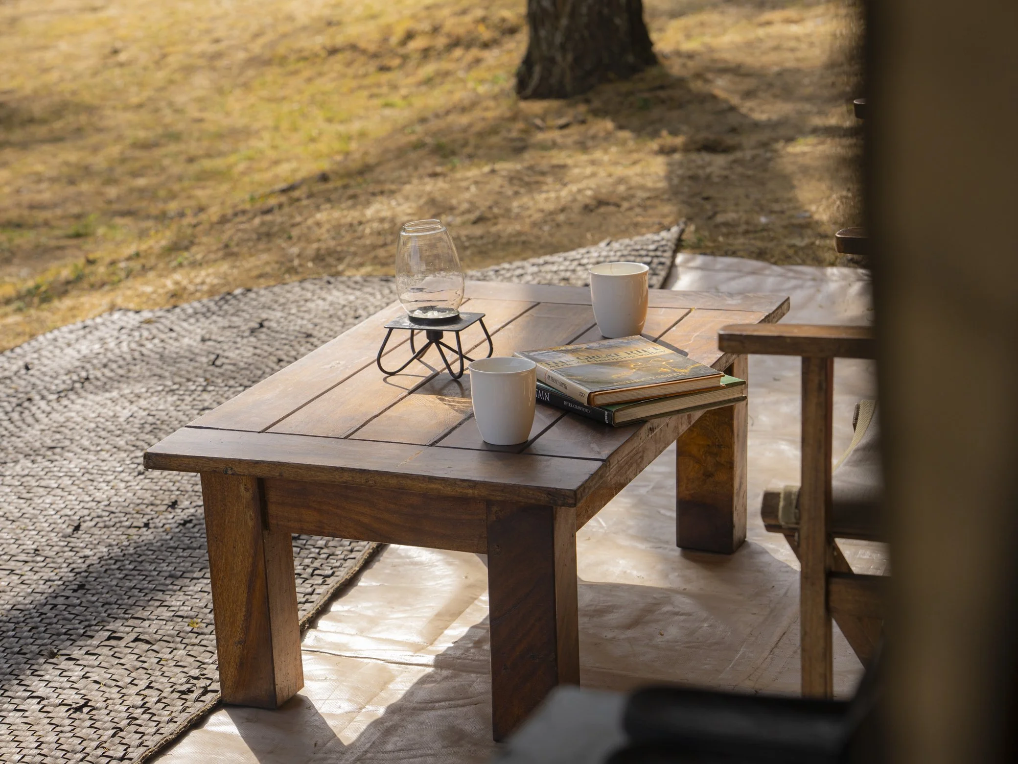 A wooden outdoor table with two white cups, a glass on a black stand, and a stack of books, set in a natural environment with trees and a grassy area.