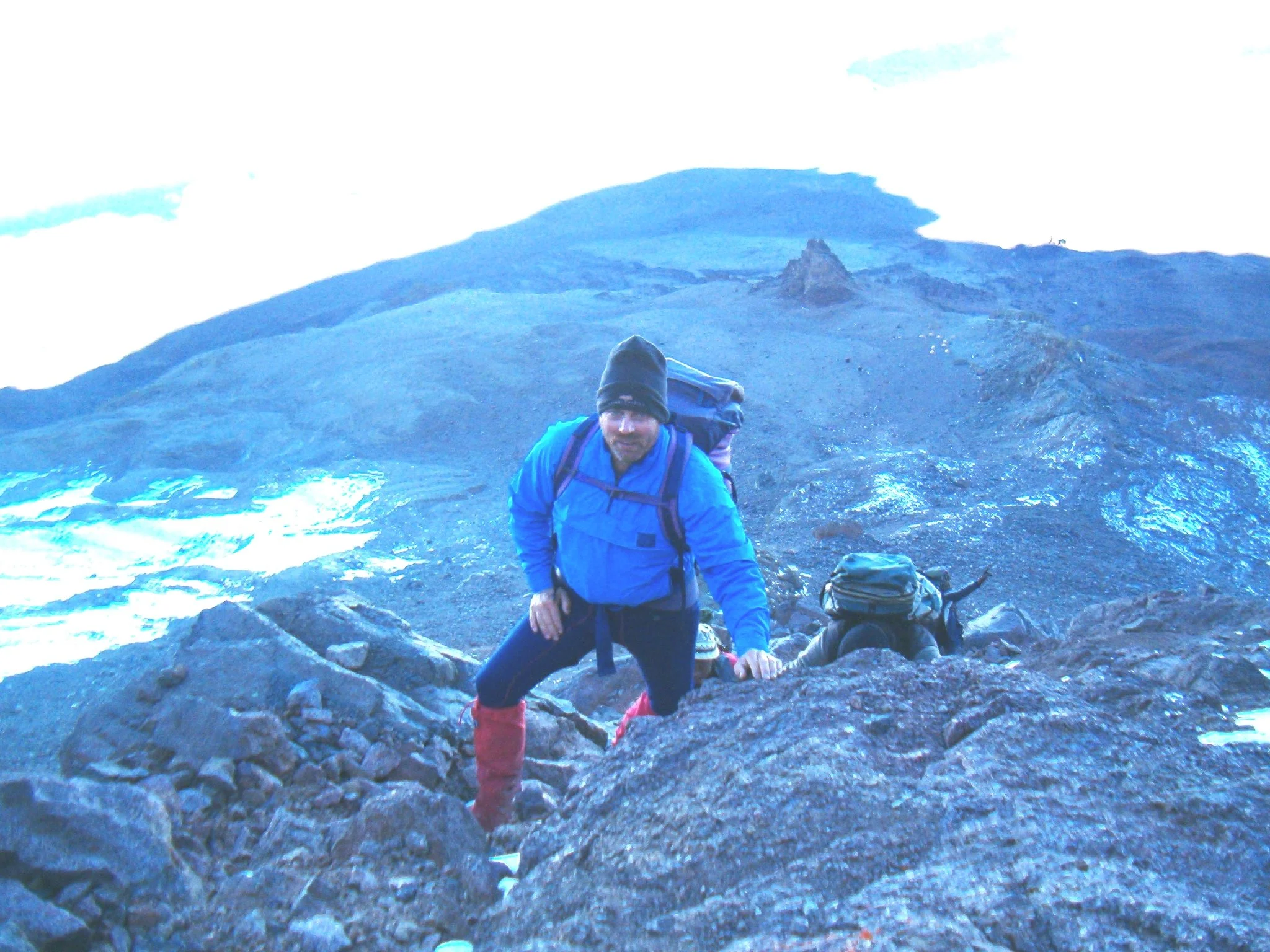 A person in a blue jacket, black beanie, and red boots hiking on a rocky, volcanic landscape with mountain peaks and a cloudy sky in the background.