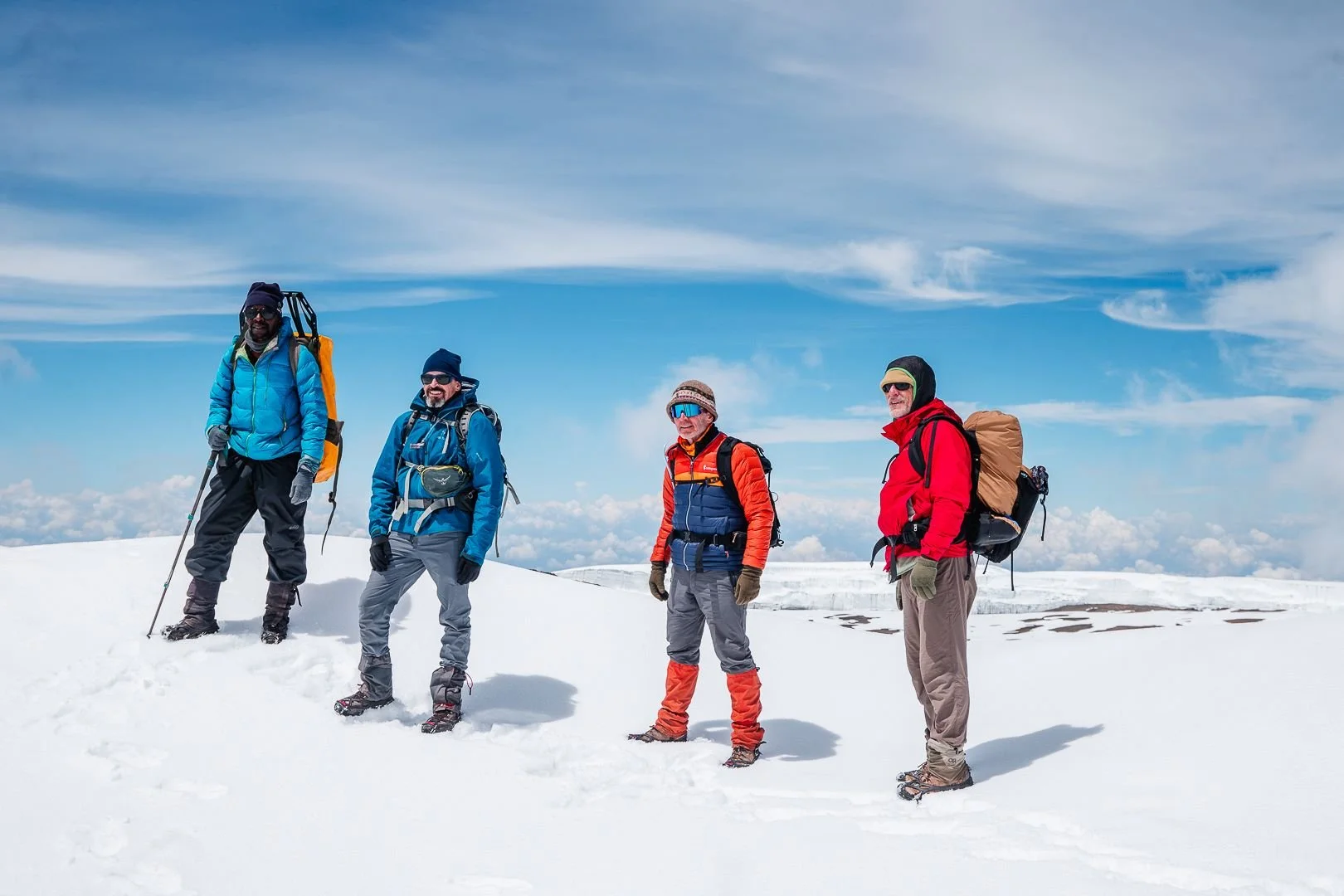 Four people dressed in winter gear and snowshoes standing on snow-covered landscape with blue sky and clouds.