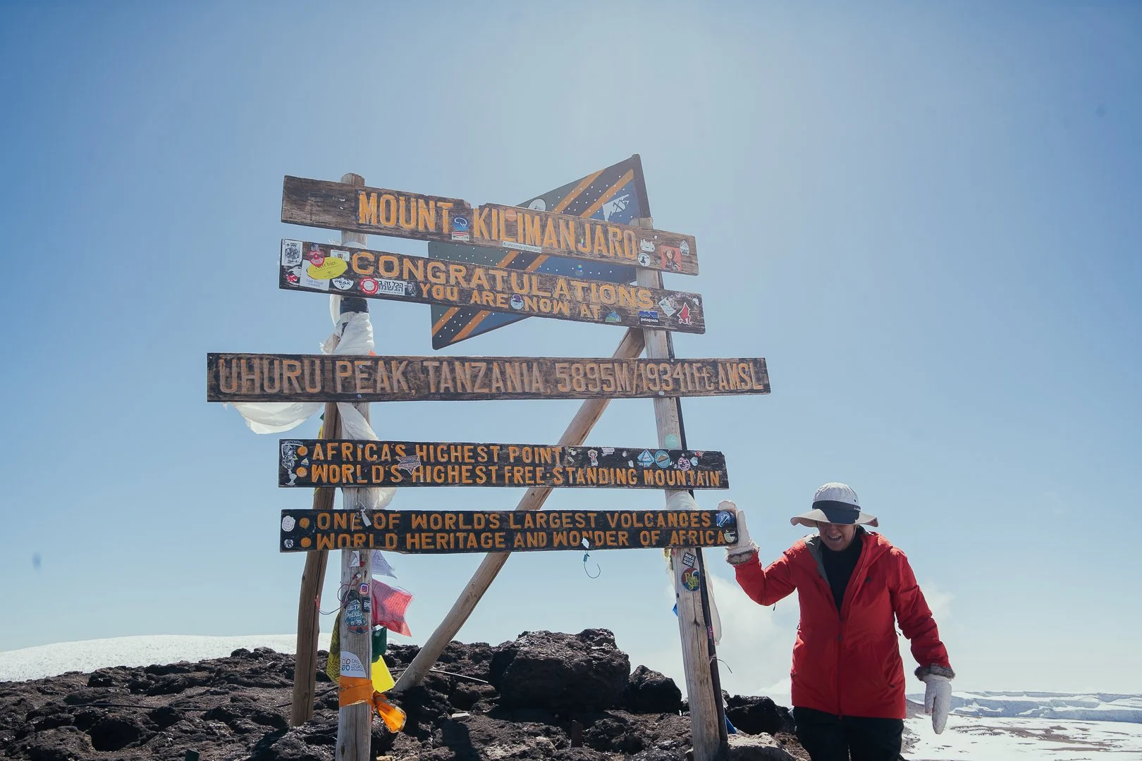 Person in red jacket and wide-brimmed hat standing next to a wooden sign at Uhuru Peak, Mount Kilimanjaro, Tanzania, showing the elevation and world records, with snow and volcanic rocks around.