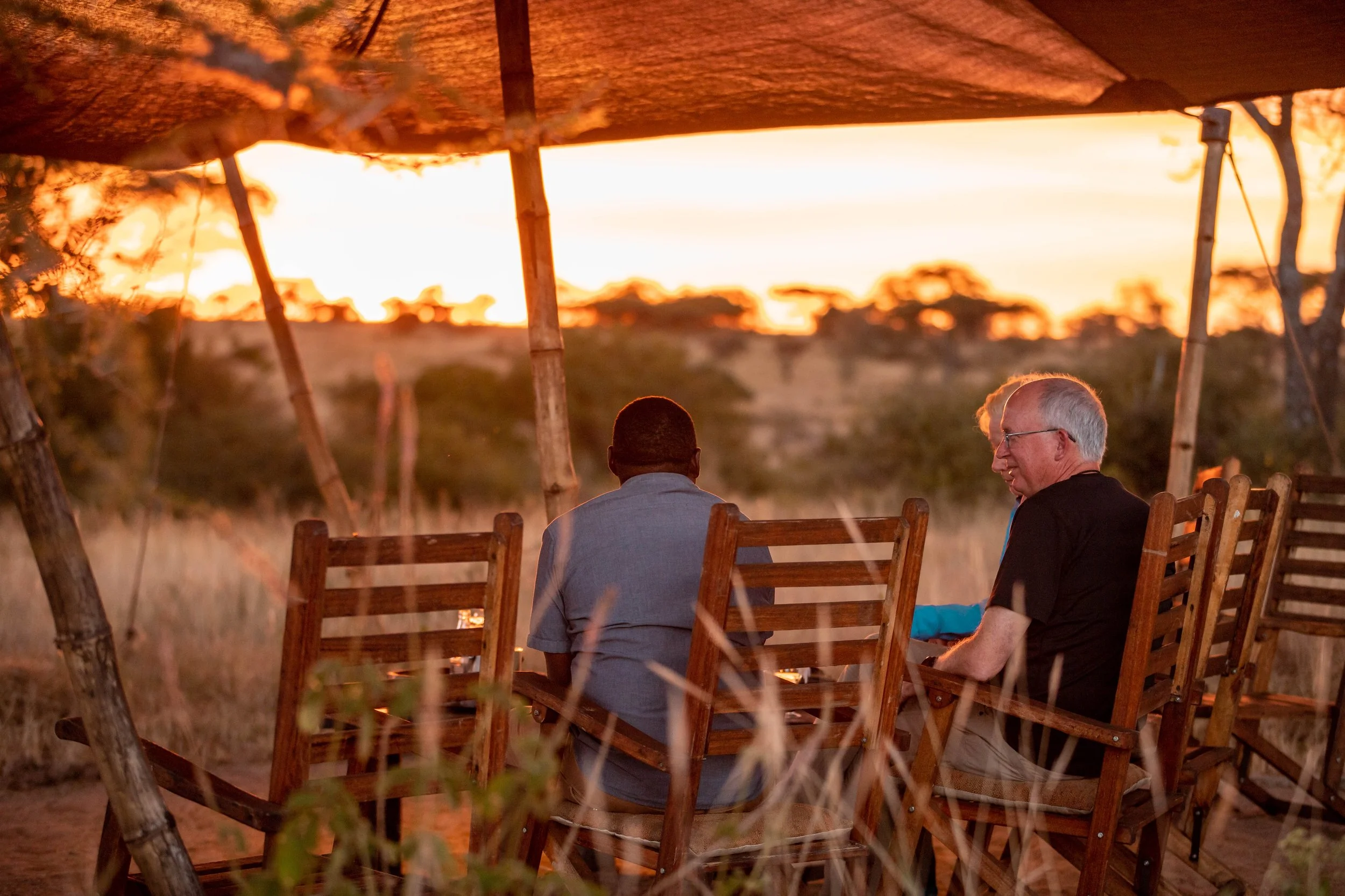 Two men sitting on wooden chairs outdoors during sunset, with a large orange sky and trees in the background.