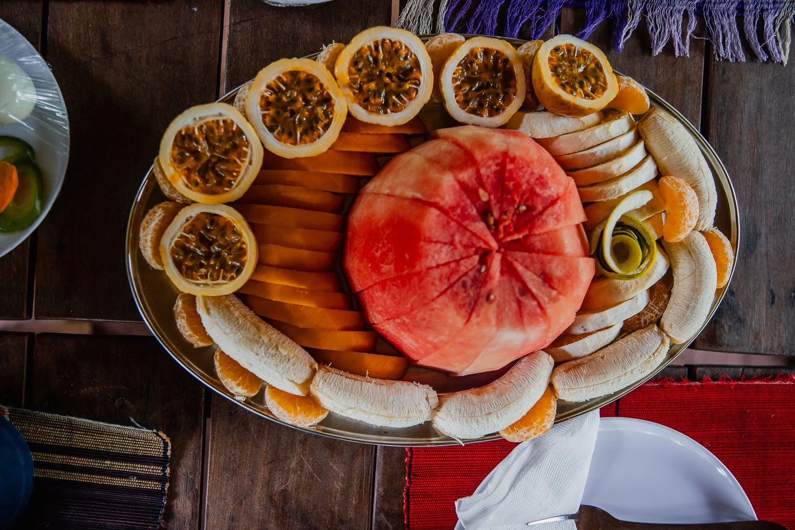 A large oval platter filled with sliced fruits, including orange, banana, passion fruit, watermelon, and a rolled cucumber on a wooden table.