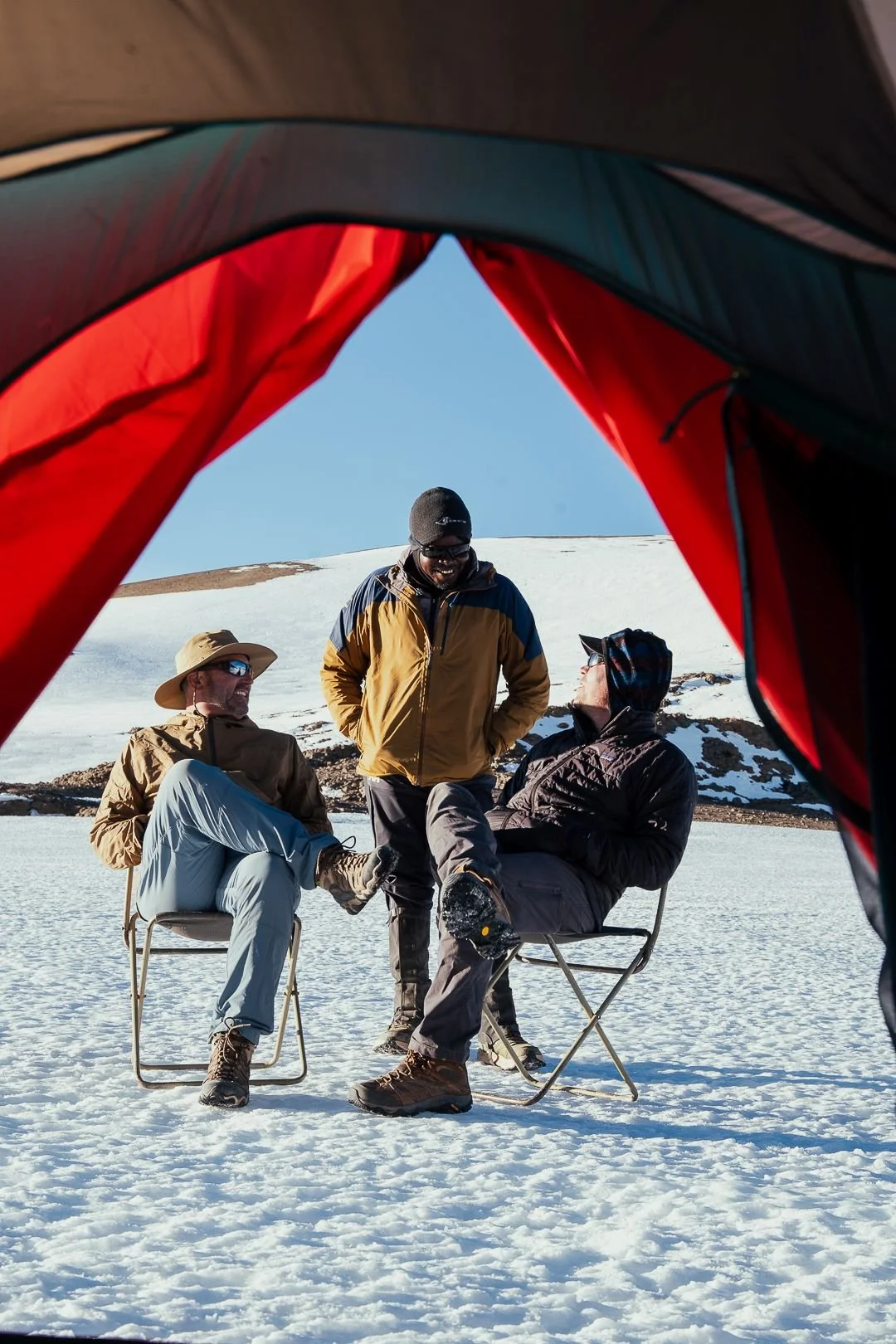 Three friends sitting and standing on snow under a bright blue sky, seen from inside a tent with red and black fabric, in a snowy landscape.