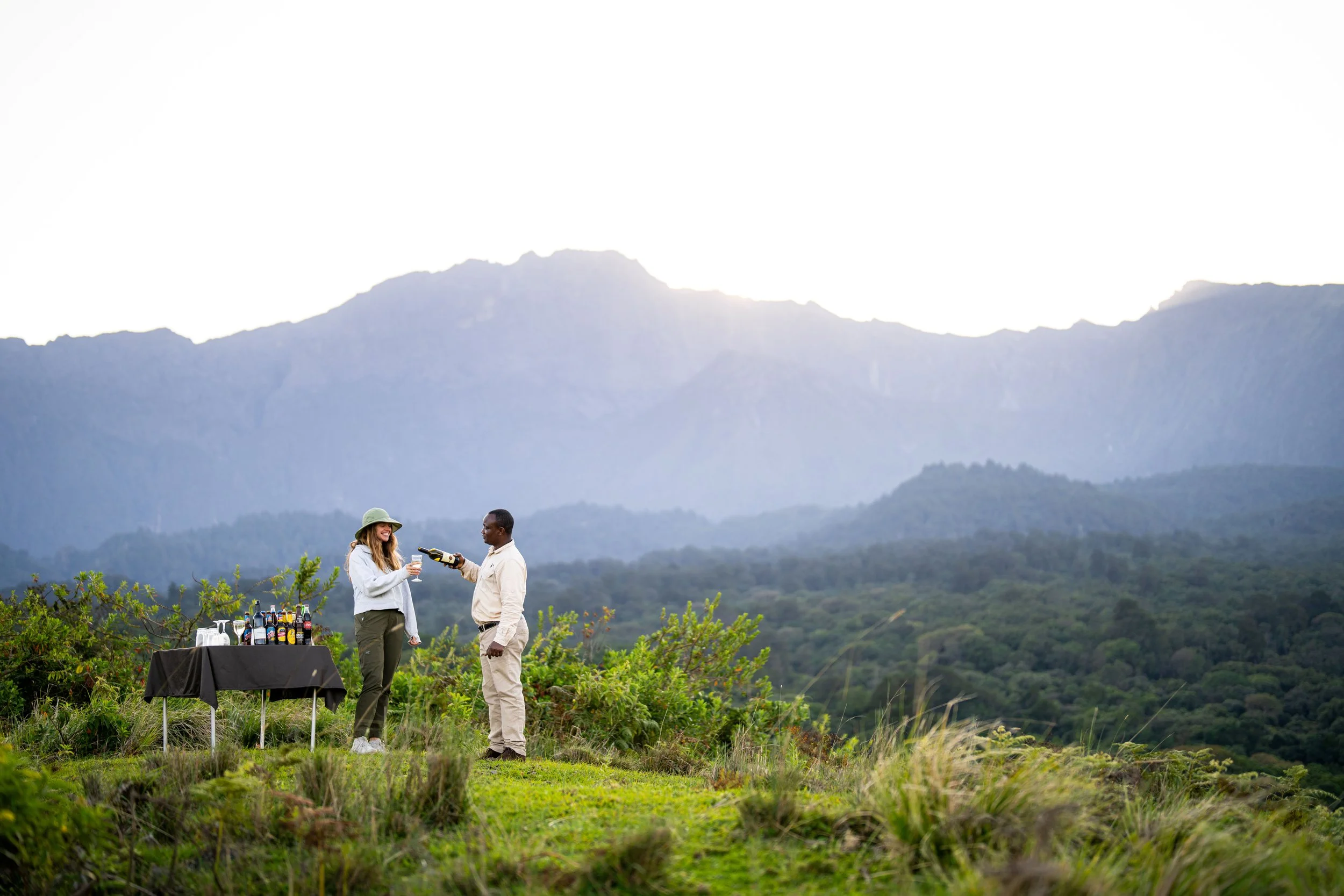 A woman in a hat and a man in beige pants having a conversation at an outdoor gathering with a table of drinks, set against a backdrop of mountains and greenery.