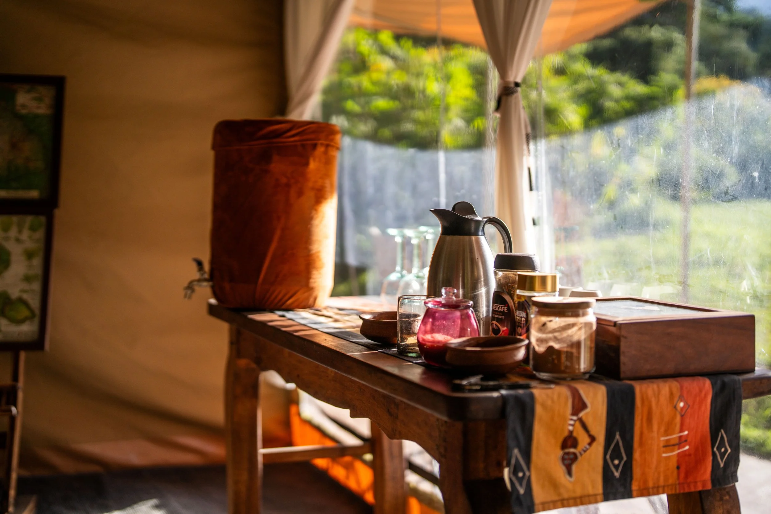 A cozy kitchen interior with a wooden countertop holding various jars, a metal kettle, and a ceramic pitcher, with a large window overlooking greenery and sunlight streaming in.