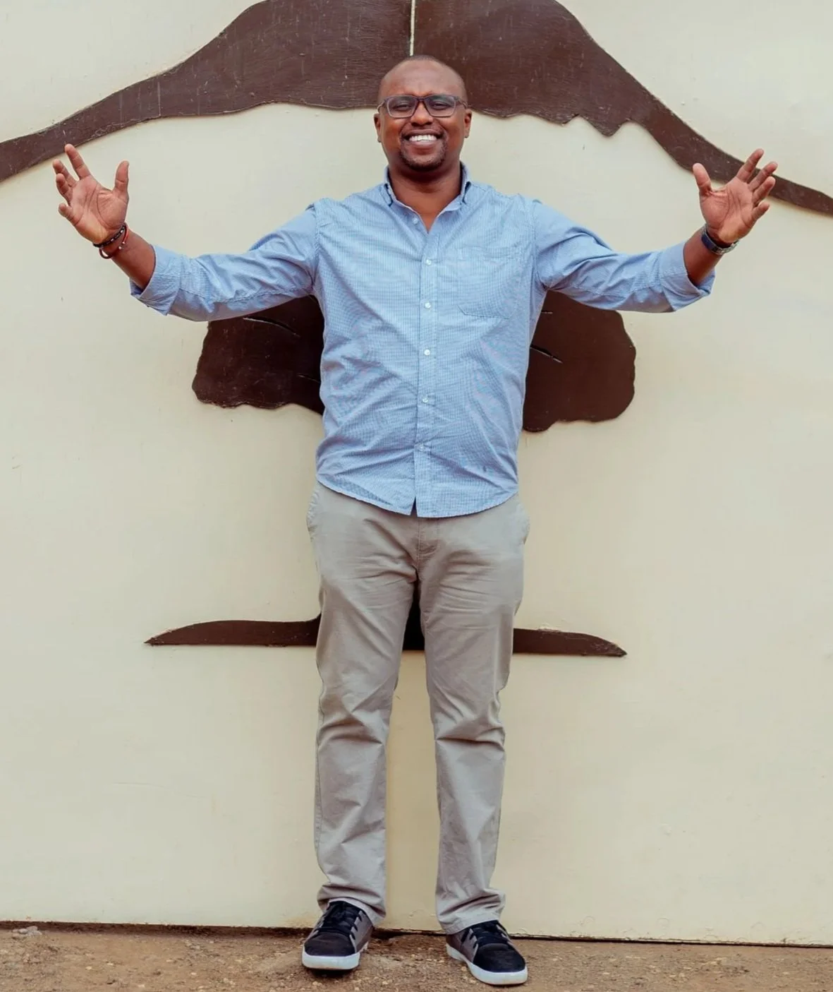 A smiling man with glasses, wearing a light blue button-up shirt and khaki pants, stands with arms outstretched in front of a wall with a brown decorative element.