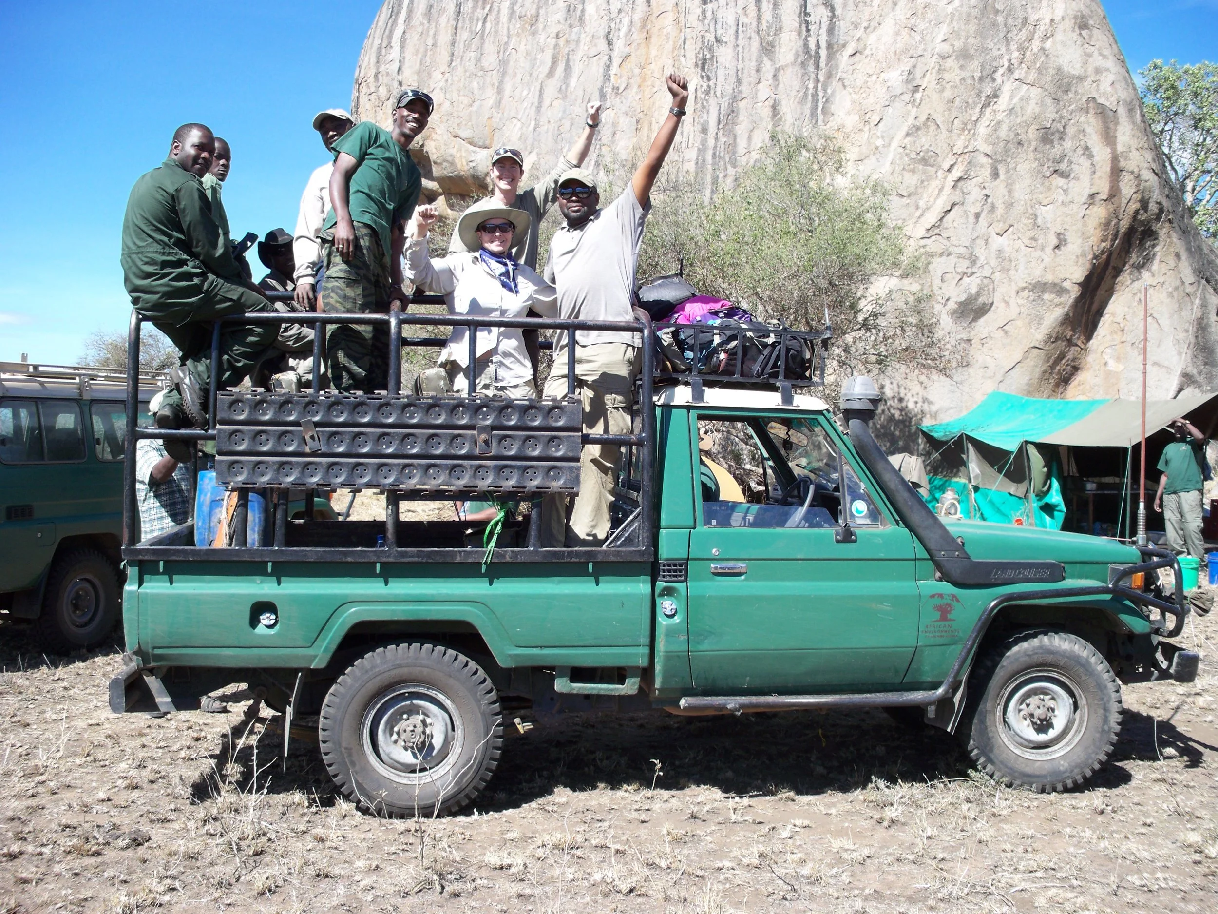 Group of six people riding on a green off-road vehicle with a large rock formation and tents in the background.