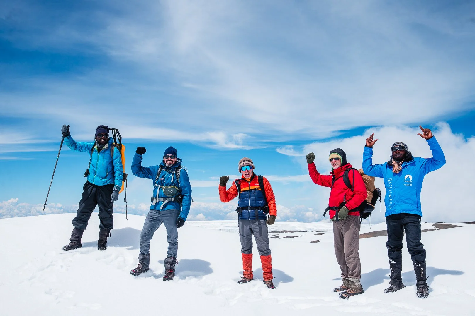 Five hikers dressed in winter gear standing on snow-covered mountain peak with blue sky and clouds in the background, raising their arms in triumph.