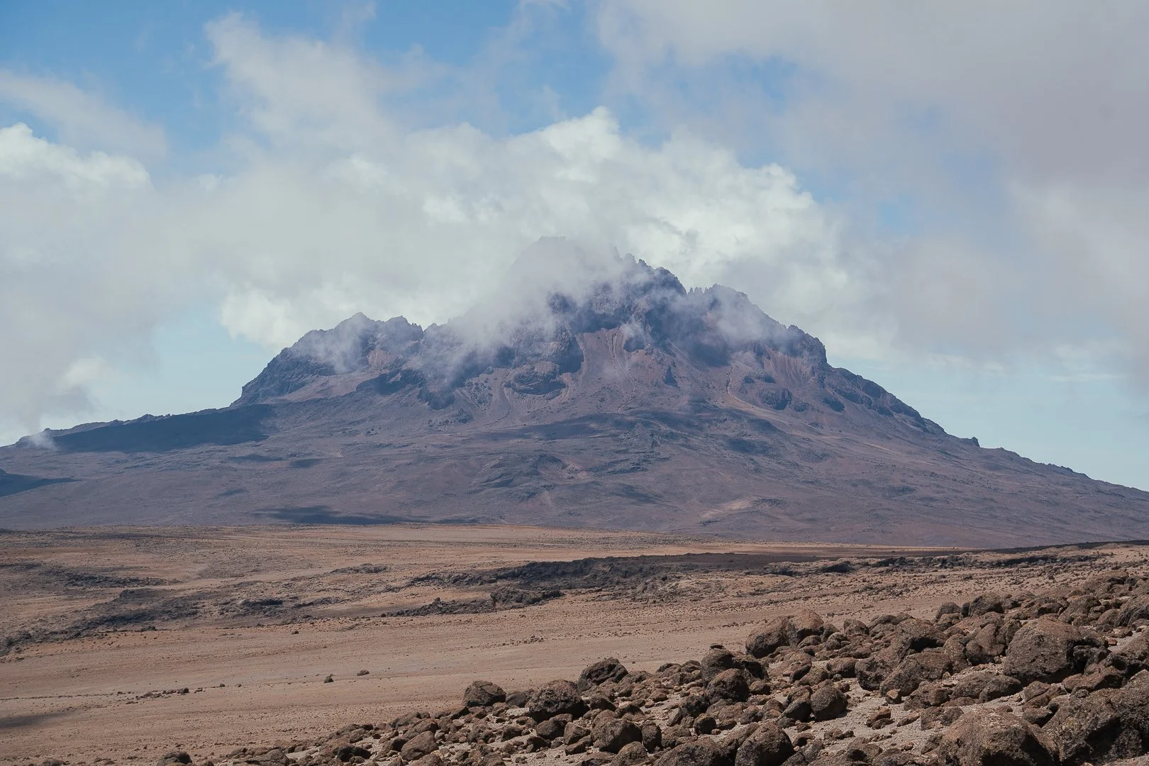 A barren desert landscape with rocky terrain in the foreground and a large mountain with some clouds around its peaks in the background.