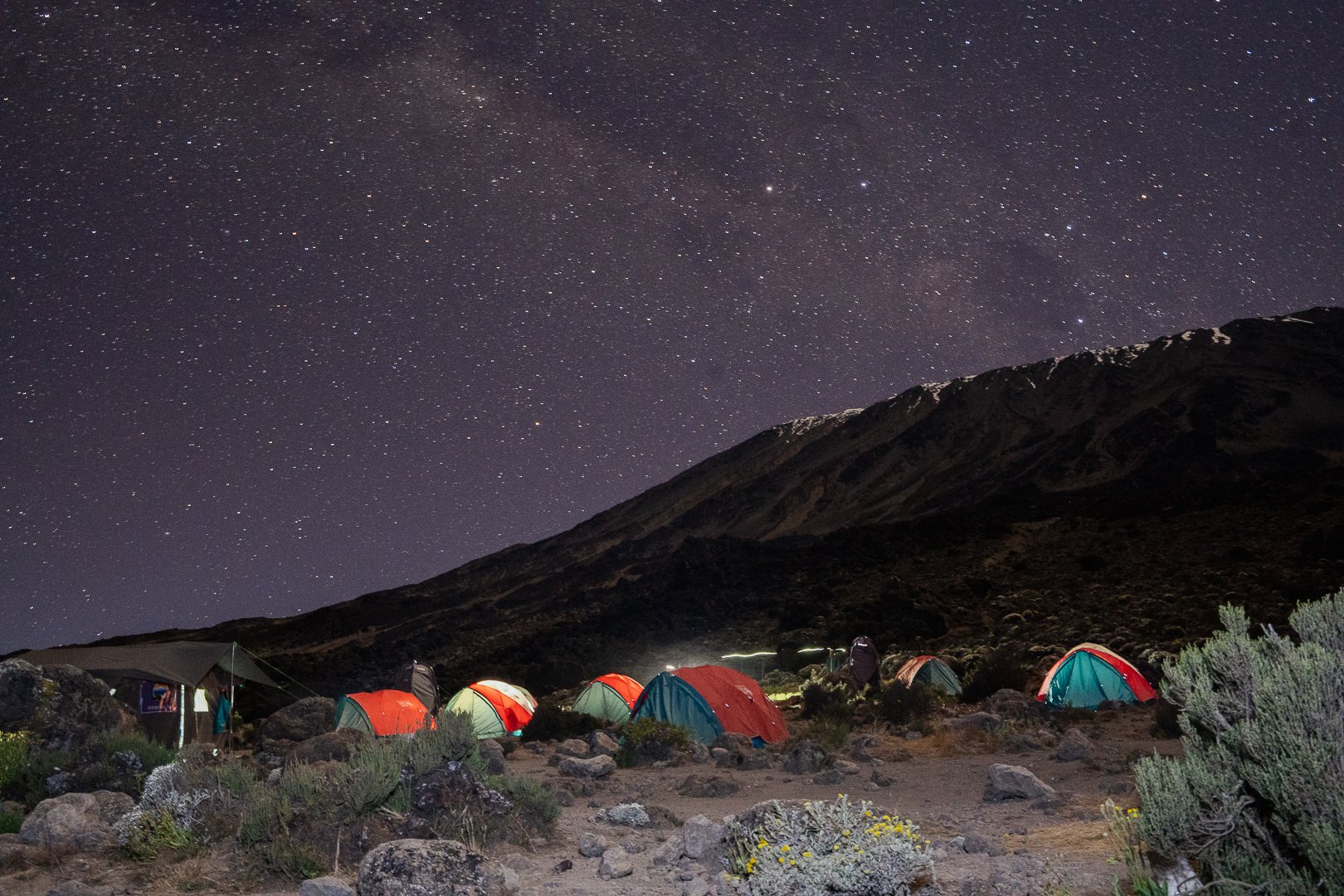 Nighttime scene of a camping site with multiple tents set up near a mountain, under a starry sky.