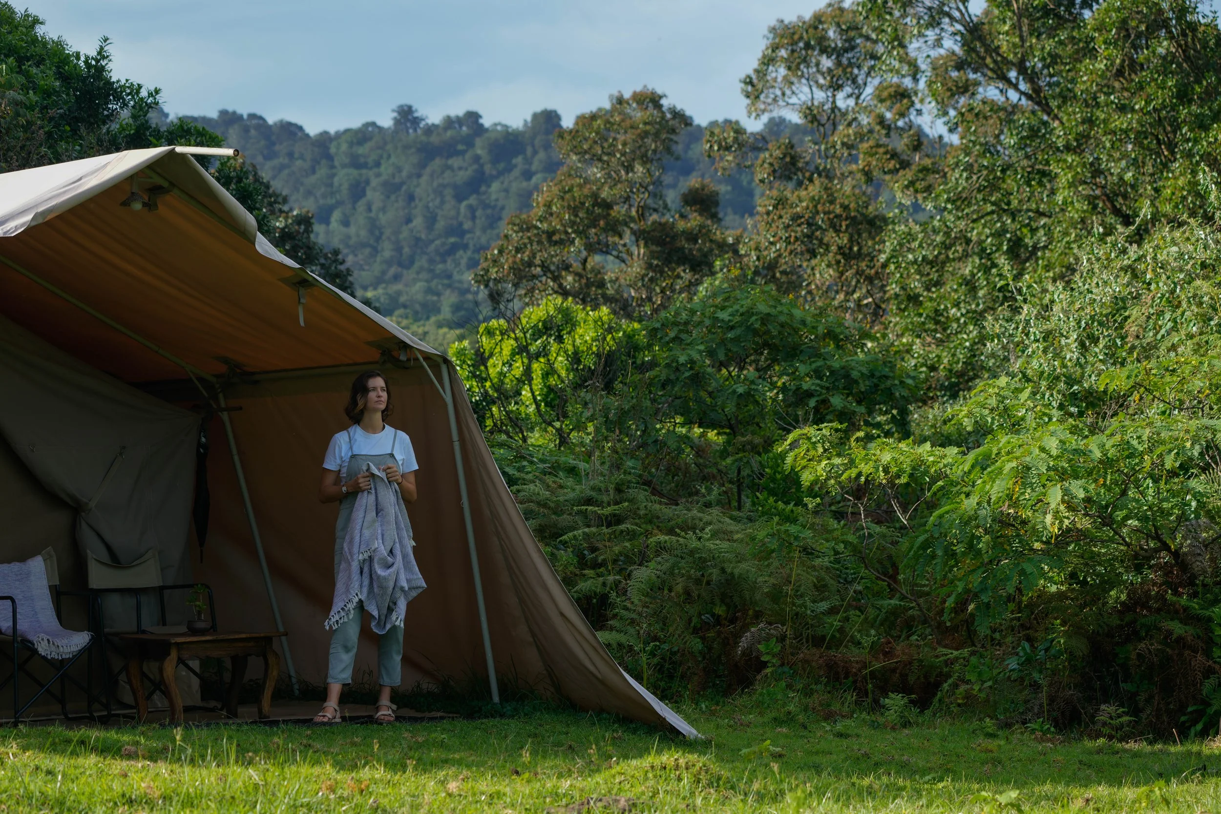 A young woman standing in front of a large camping tent in a lush green forested area, holding a towel, with trees and a hill in the background.