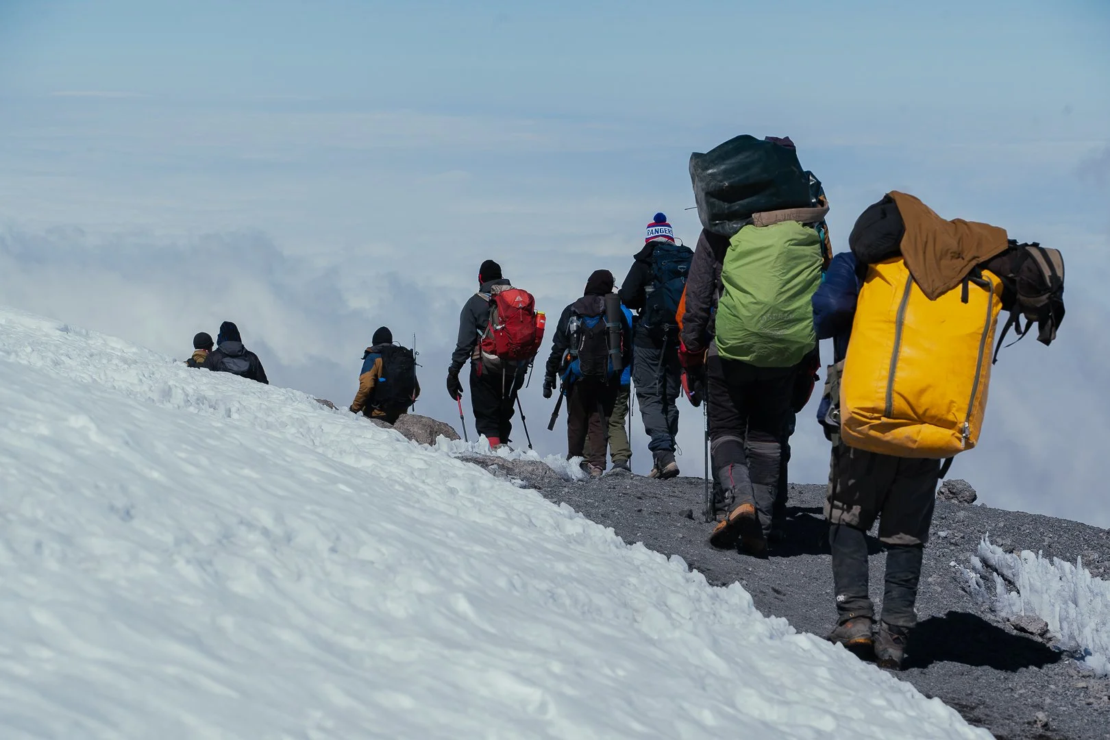 Group of hikers ascending a snowy mountain trail with backpacks and trekking poles.