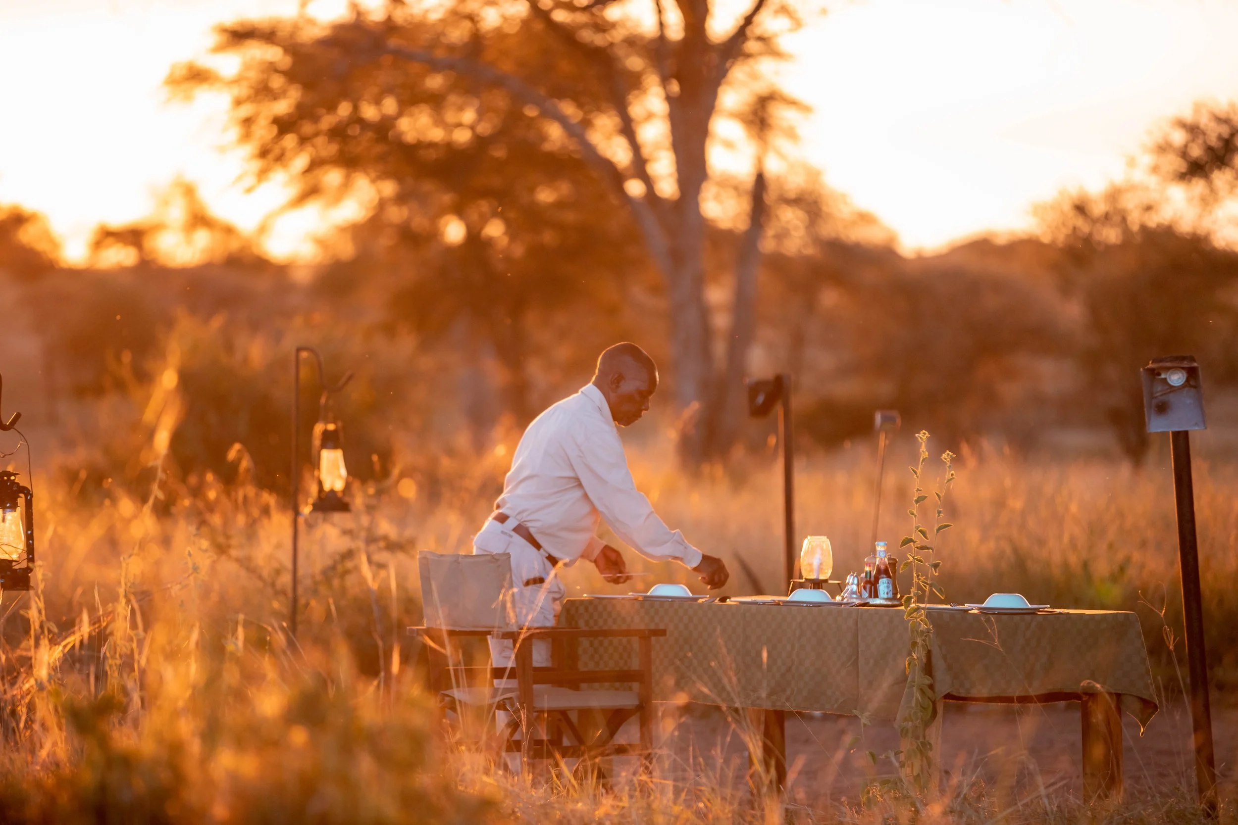A person dressed in white preparing food outdoors during sunset, with trees and decorative lamps in the background.