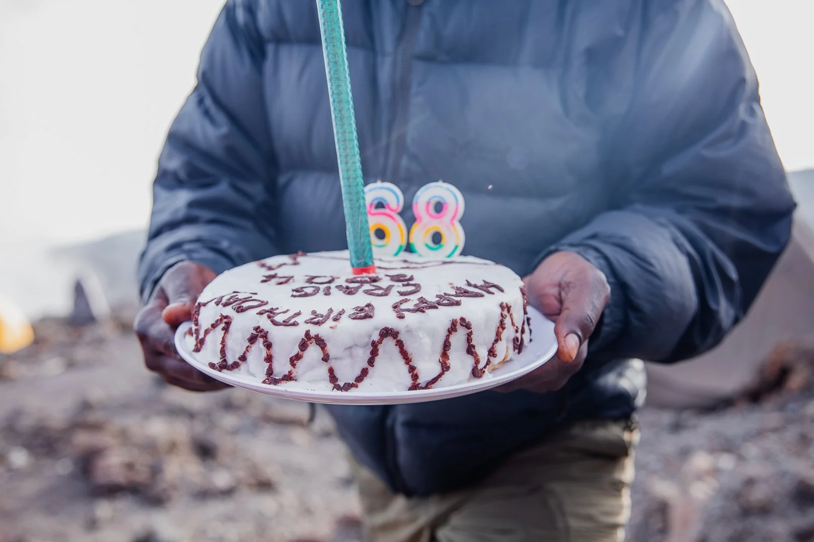 Person in a dark jacket holding a birthday cake with candles shaped as the number 88, topped with colorful number candles and chocolate writing, outdoors near a body of water.