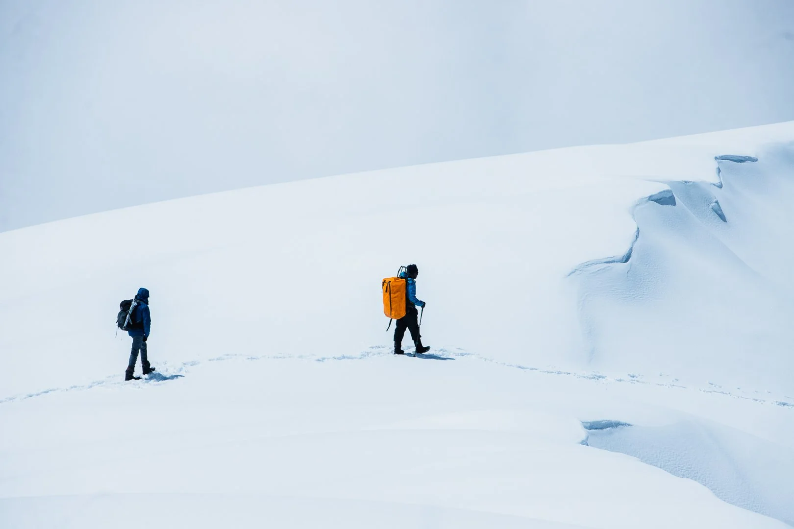 Two mountaineers dressed in winter gear walking through snow on a mountain, with snow-covered peaks in the background.