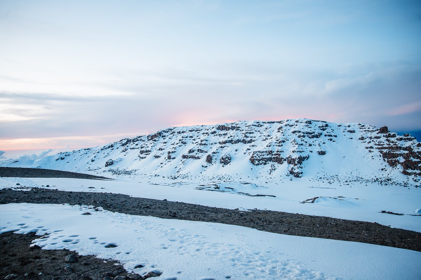 Snow-covered mountain with a rocky base, under a cloudy sky at sunset or sunrise.