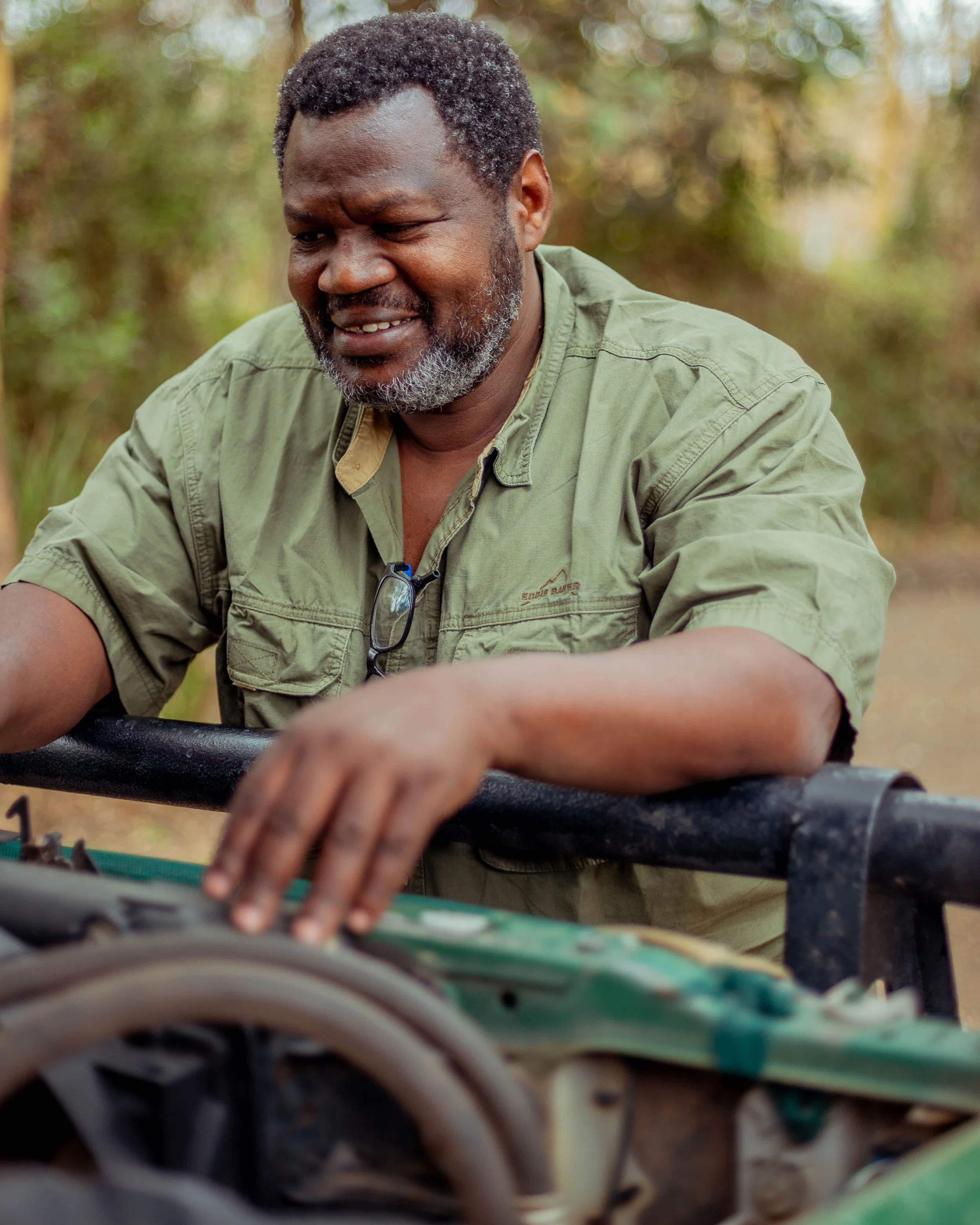 A man with a beard and short curly hair, wearing a green shirt and glasses hanging from his neck, outdoors in a wooded area, looking down and smiling slightly.