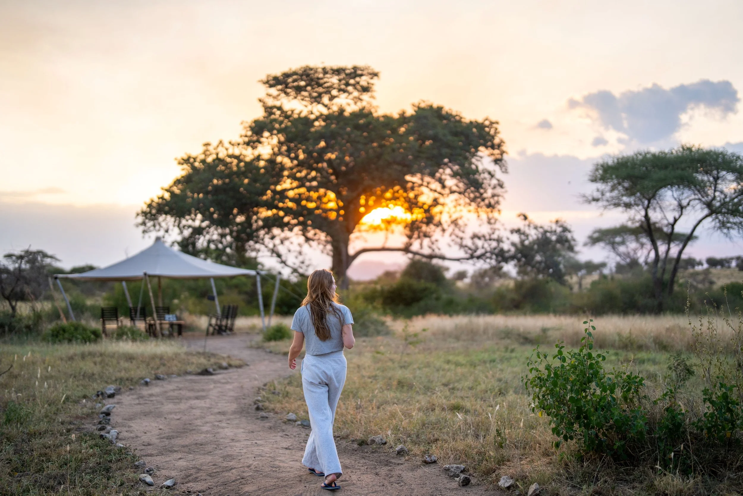 A young woman with long hair wearing a gray t-shirt and white pants walking on a dirt path in a rural area during sunset. There are trees, a tent, and outdoor furniture in the background, creating a peaceful outdoor scene.