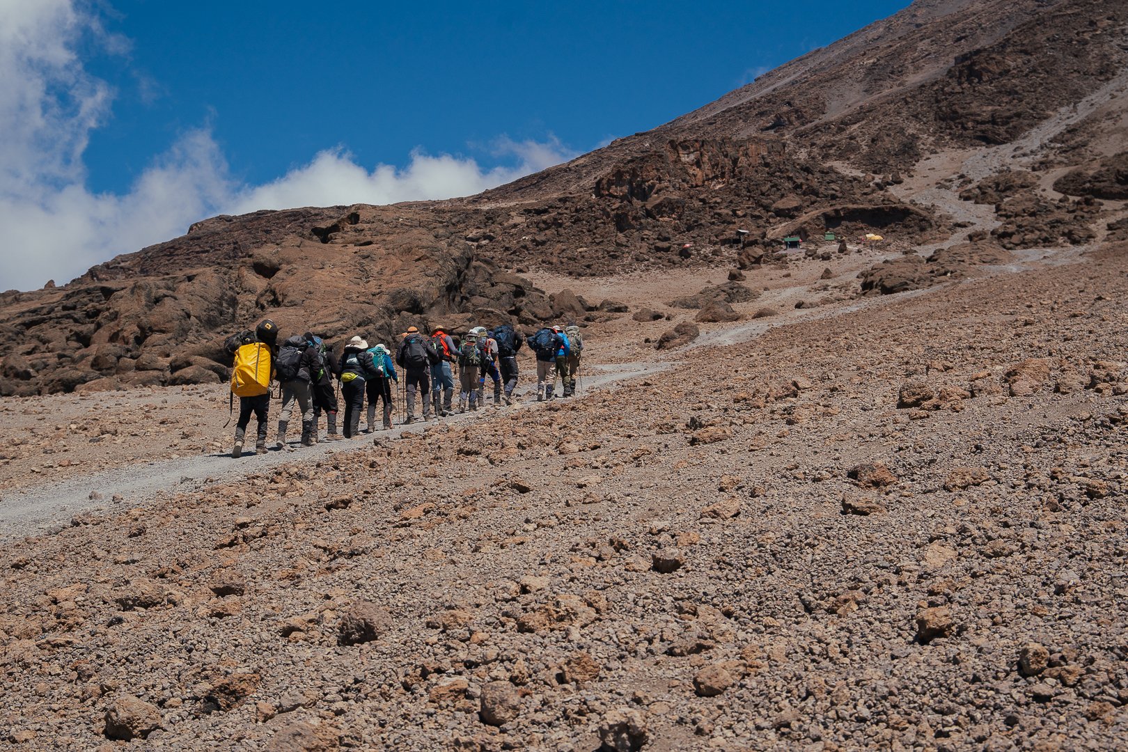 Group of hikers walking on a rocky mountain trail with brown terrain and a blue sky.
