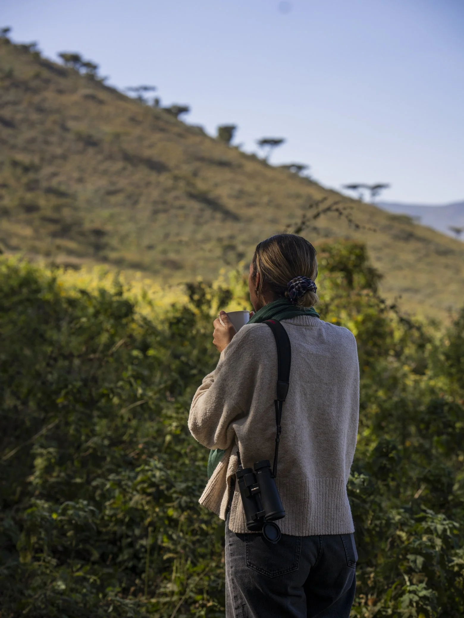 A person with brown hair tied in a bun, wearing a beige sweater and black pants, holding a white mug, and looking at a hillside with scattered trees, a clear blue sky, and lush green bushes in the foreground.