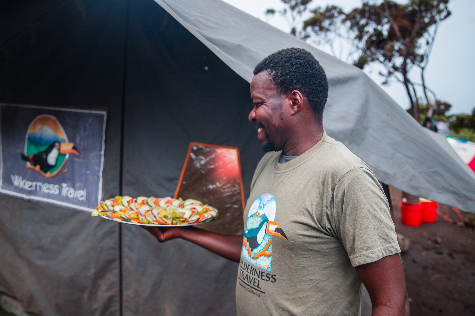 A man smiling and holding a plate of food outdoors near a tent with a 'Wilderness Travel' sign.