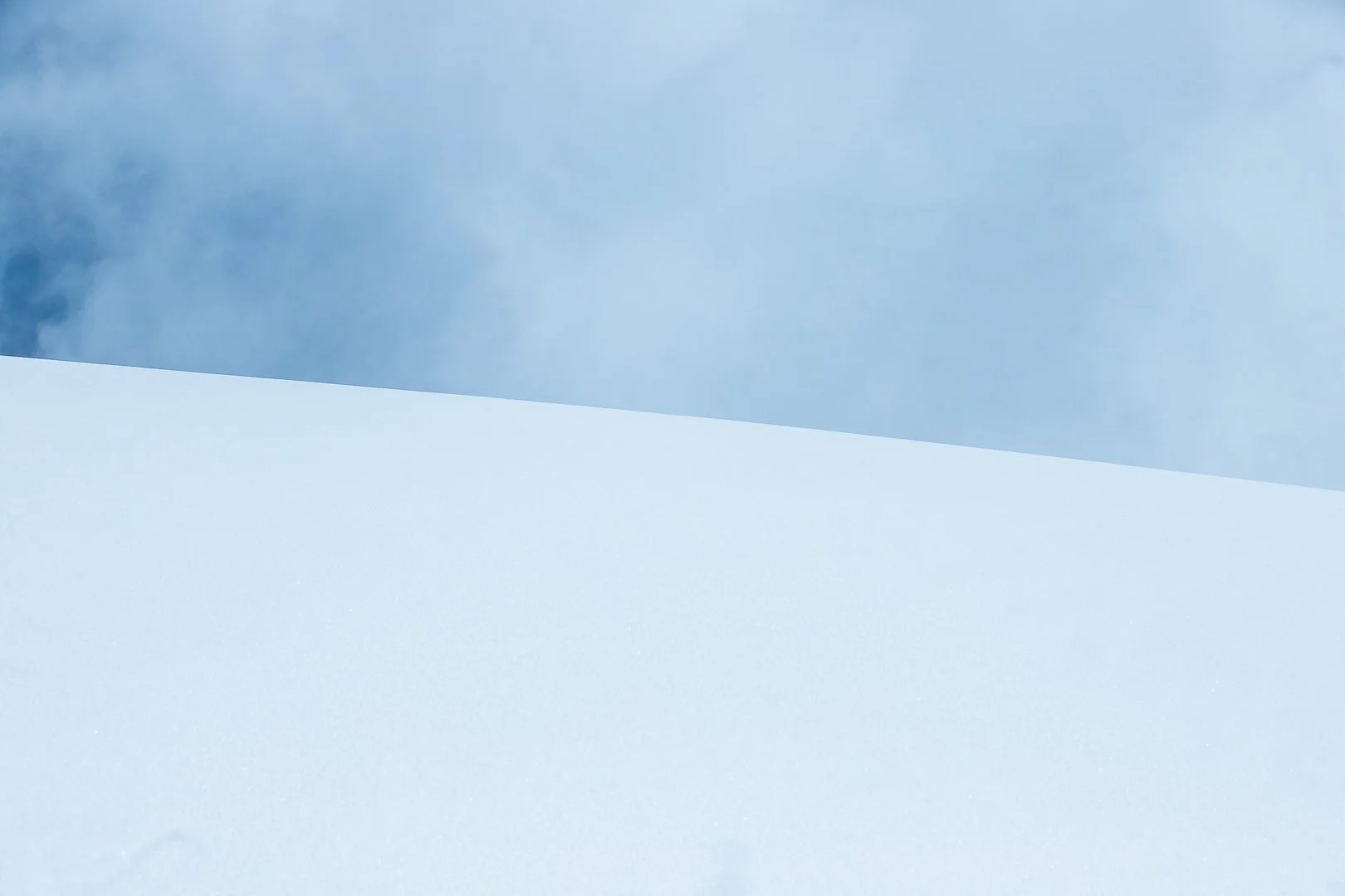 A landscape scene featuring a cloudy sky with some blue visible, and a large, smooth, white snow-covered surface in the foreground.