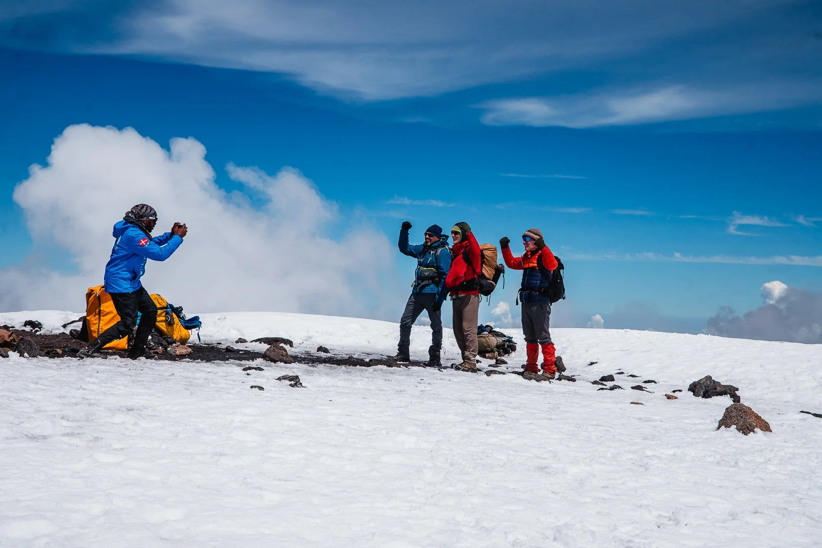Four hikers in winter gear on snowy terrain, with one person taking a photo of the other three, who are posing with raised fists under a blue sky with clouds.