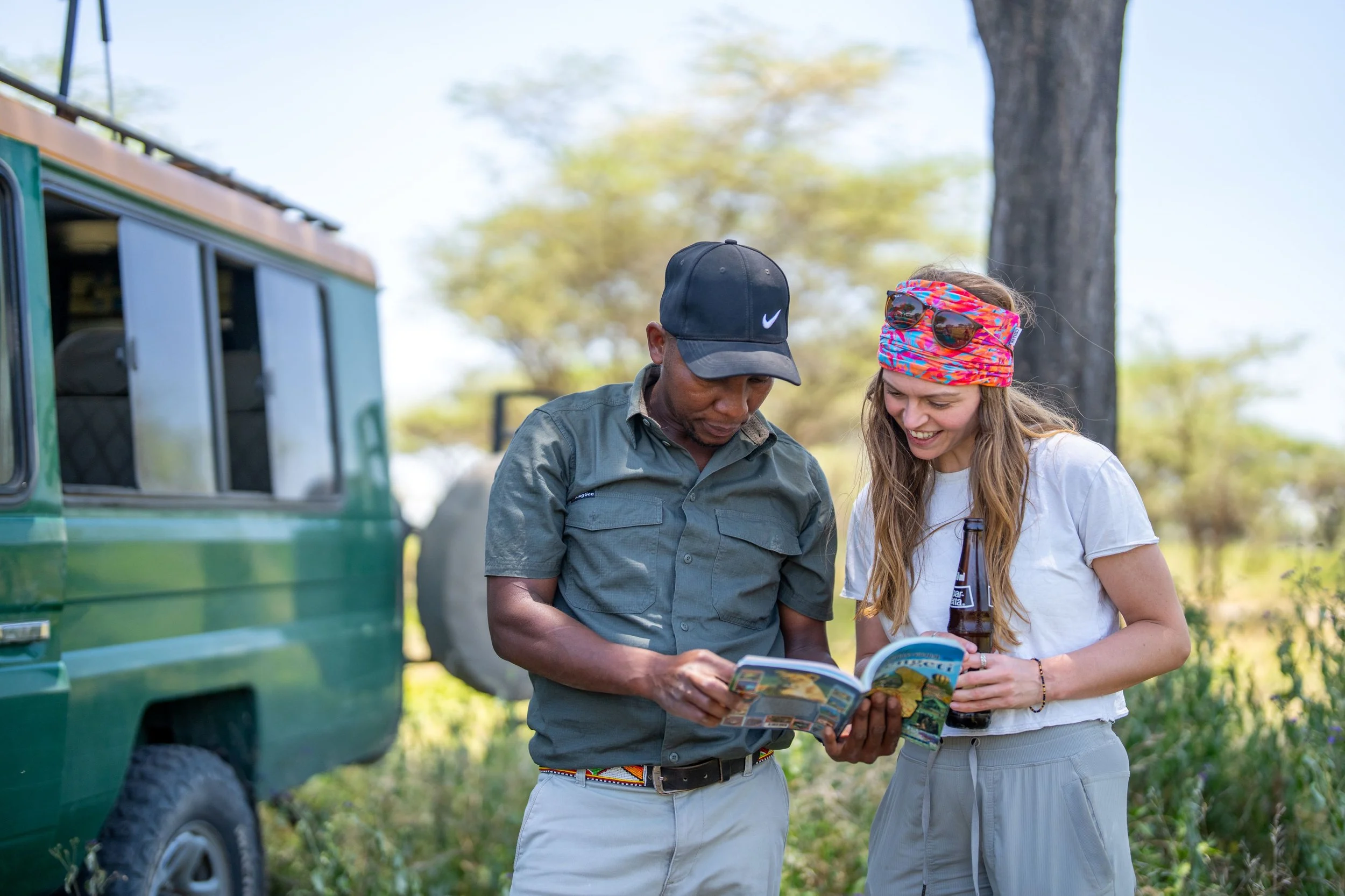 Two people, a man and a woman, are outdoors near a green vehicle, looking at a map or brochure together. The man is wearing a dark baseball cap and a green shirt, and the woman is wearing sunglasses on her head, a colorful headband, and a white t-shirt. It appears to be a sunny day with trees in the background.