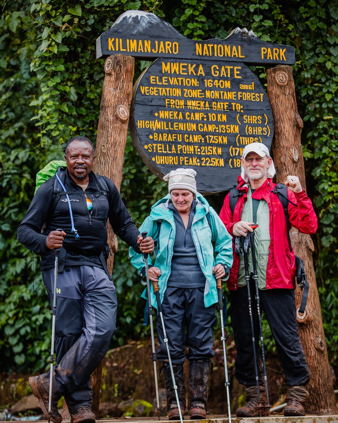 Three hikers with backpacks and trekking poles standing in front of a wooden sign at Kilimanjaro National Park. The sign provides elevation and distance details for various points on Mount Kilimanjaro, including Mweka Gate, Mweka Camp, Millennium Cam