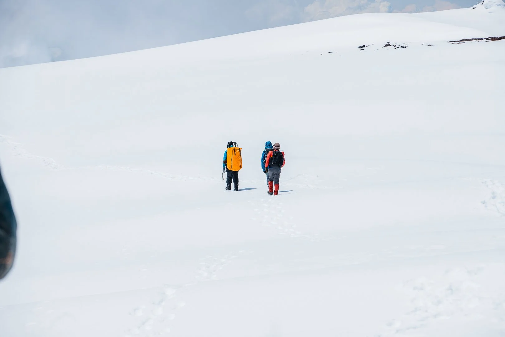 Three people in winter gear walking through a snow-covered landscape, with snowy hills in the background.