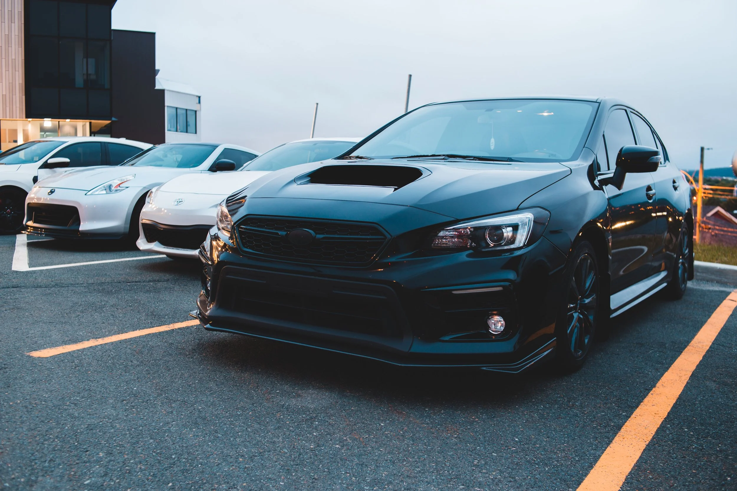 Several cars parked in a parking lot with a modern building in the background, under an overcast sky.