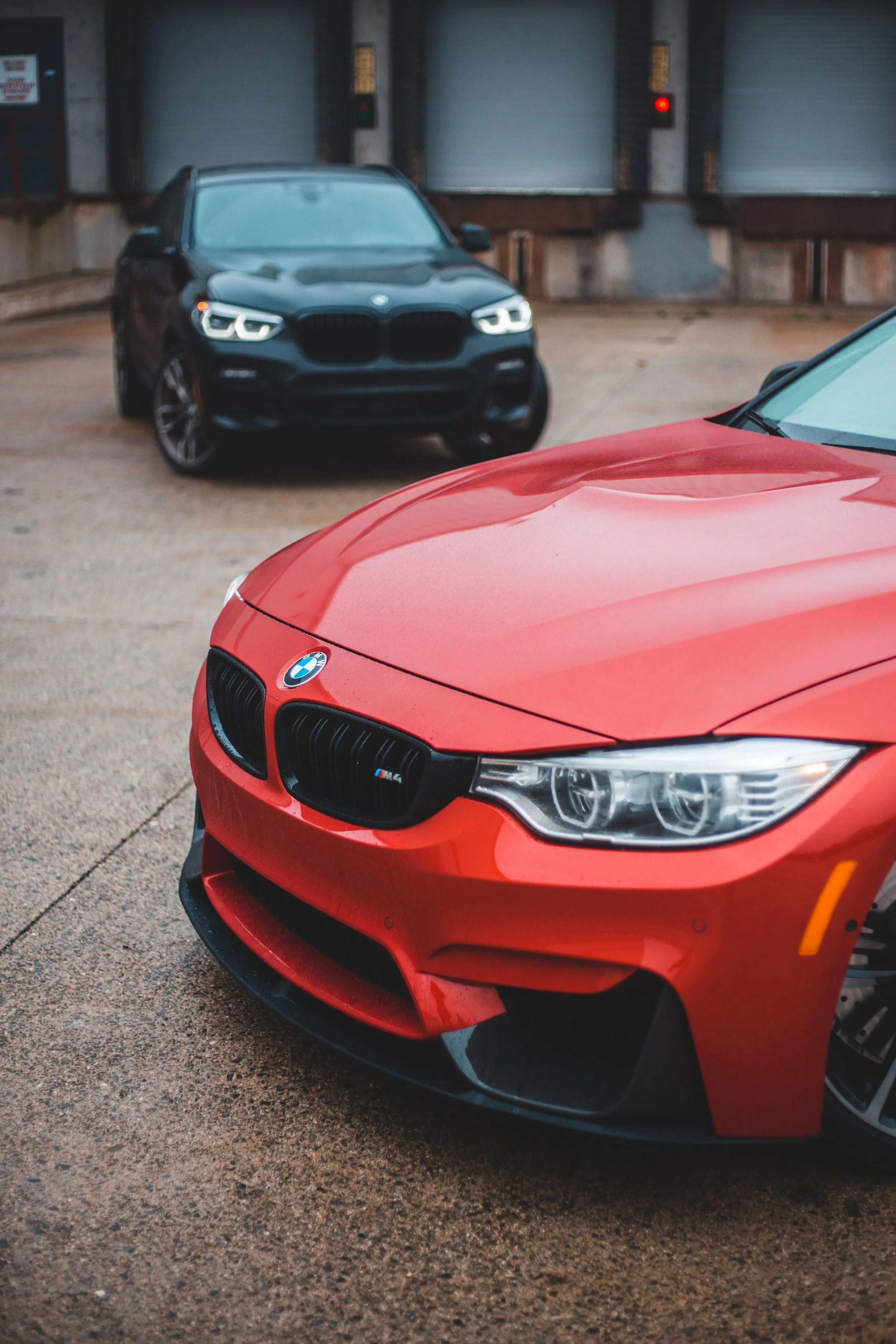 Close-up of the front of a red BMW sports car, with a black BMW SUV in the background across a parking lot near loading dock doors.