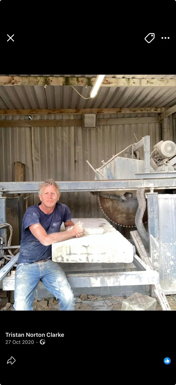 Tristan sits looking to camera he's wearing a blue top and blue jeans with his stone saw behind him