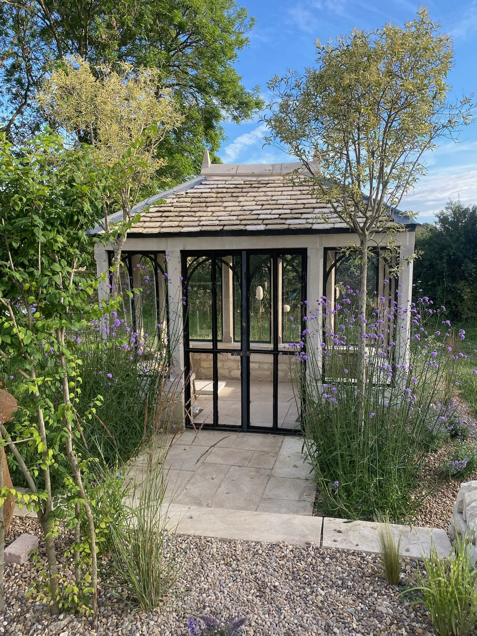 A lime stone mullion house sits nestled amongst english summer flowers, it has black framed medieval styled windows and doors and a lime stone slated roof