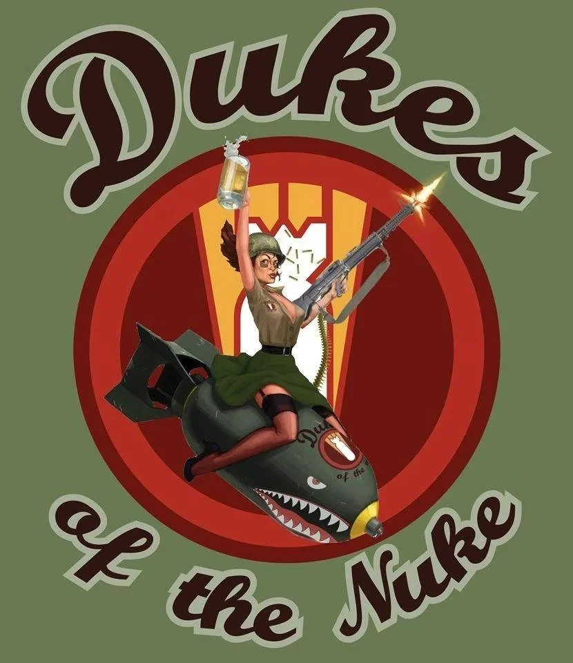 A cartoon woman in a military uniform and a helmet, riding a rocket with a shark face design. She is holding a beer mug and firing a rifle. The background features a military emblem with a red and yellow shield and the text 'Duke's of the Nuke'.