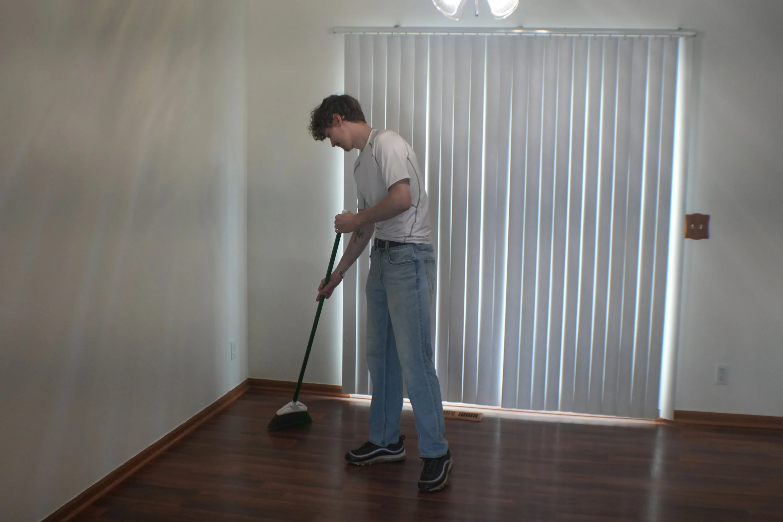 A person with curly hair wearing a white t-shirt and blue jeans is cleaning hardwood floors with a mop in a room with white walls, vertical blinds covering a sliding glass door, and a wooden floor.
