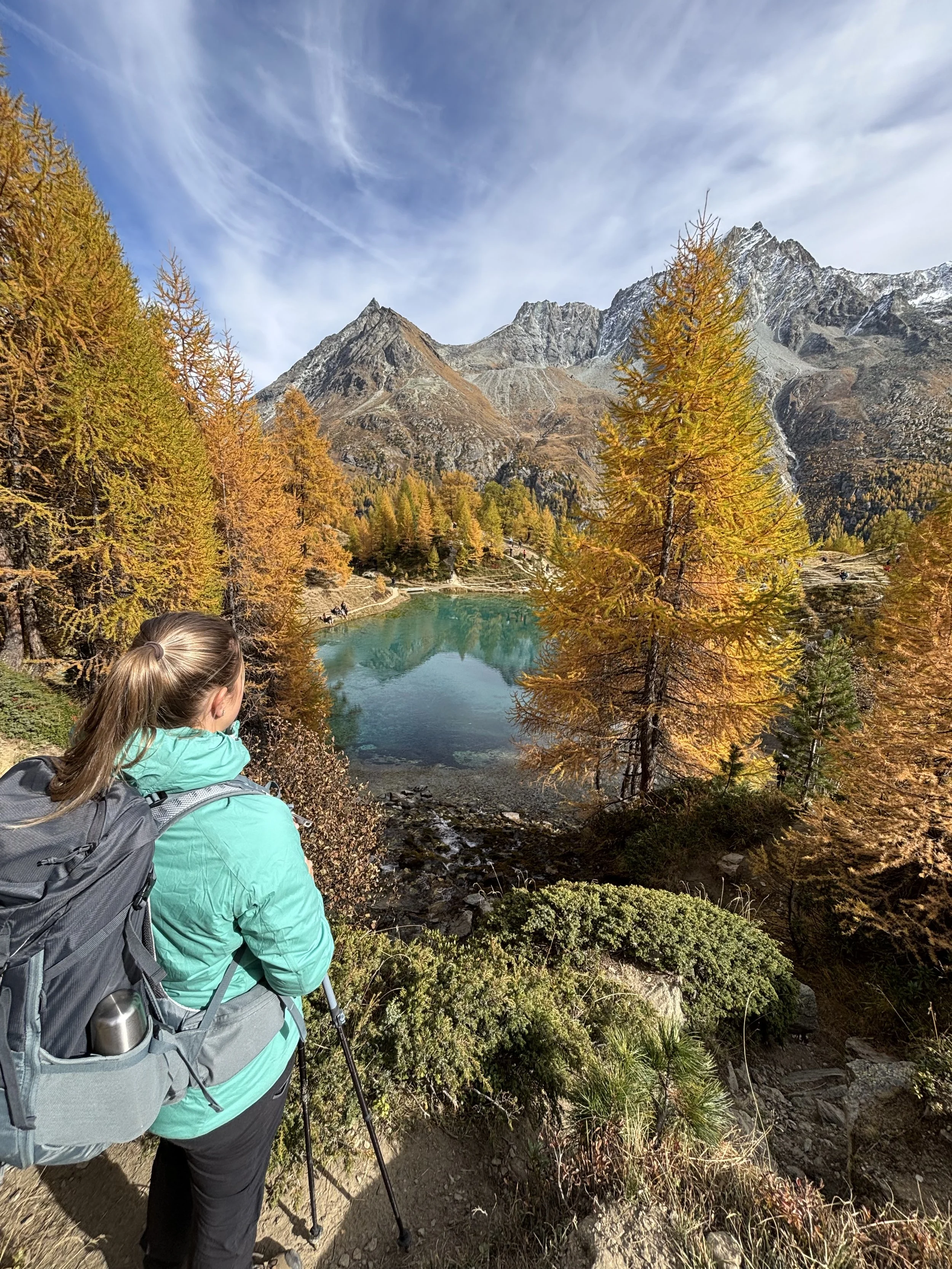 Une femme équipée d'un sac à dos et de bâtons de marche regarde un paysage montagneux avec des arbres aux couleurs automnales, un lac turquoise au premier plan, et des montagnes escarpées en arrière-plan sous un ciel partiellement nuageux.