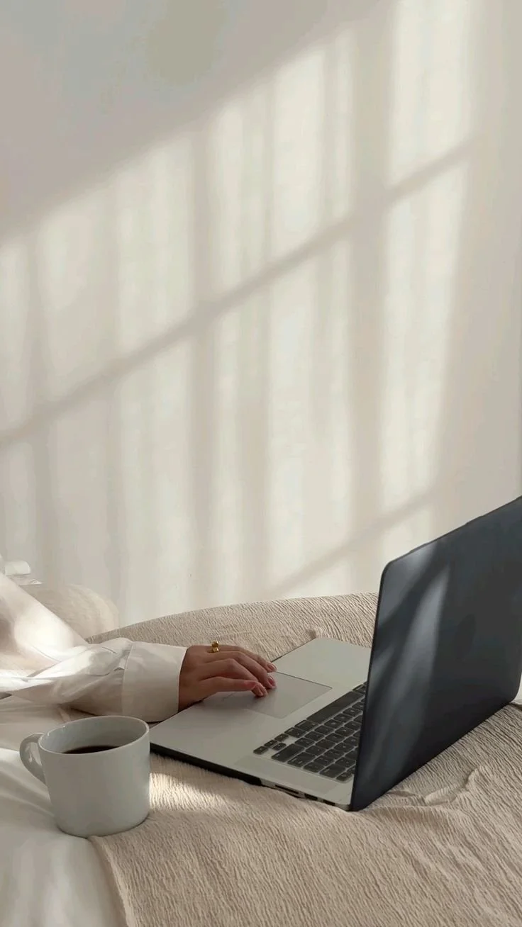 Person working on a laptop with a cup of coffee nearby, on a beige bed with sunlight casting grid-like shadows on the wall.