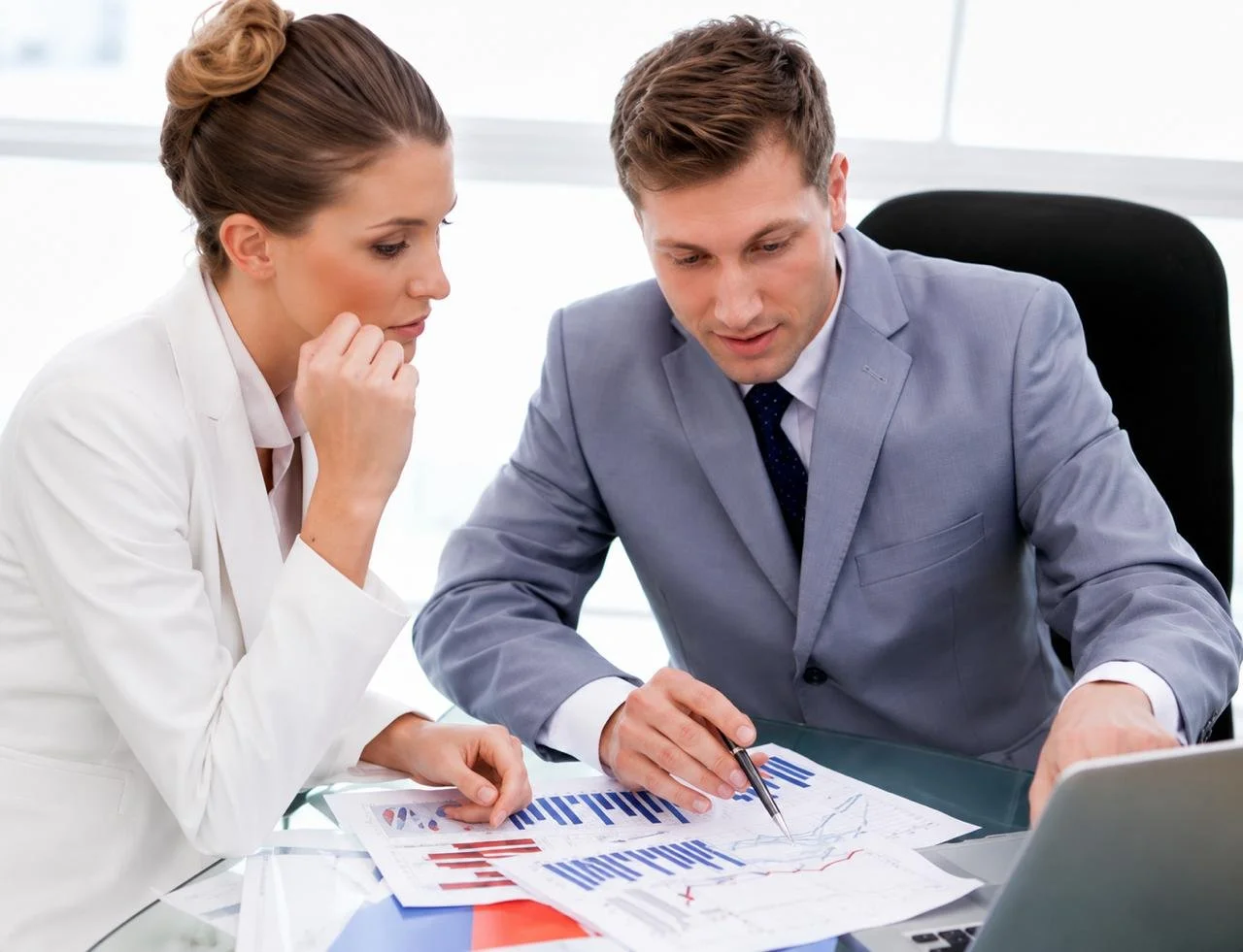 A man and woman in business attire discussing charts and graphs at a desk in an office.