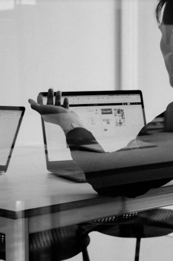 A person is sitting at a desk using a laptop, with a smartphone placed to the left on the desk. The image is in black and white.