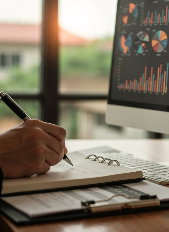 A person writing in a notebook at a desk with a computer displaying financial charts and graphs.