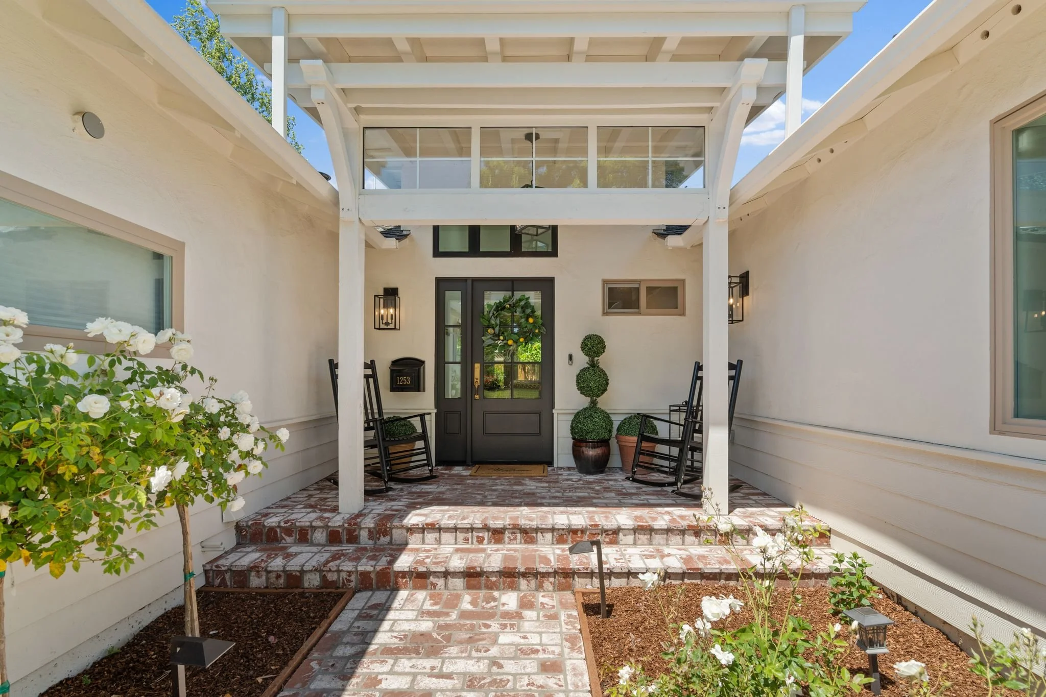 Front entrance porch of a house with brick steps, black front door decorated with a green wreath, and white siding. There are black rocking chairs, potted plants, and outdoor lighting.