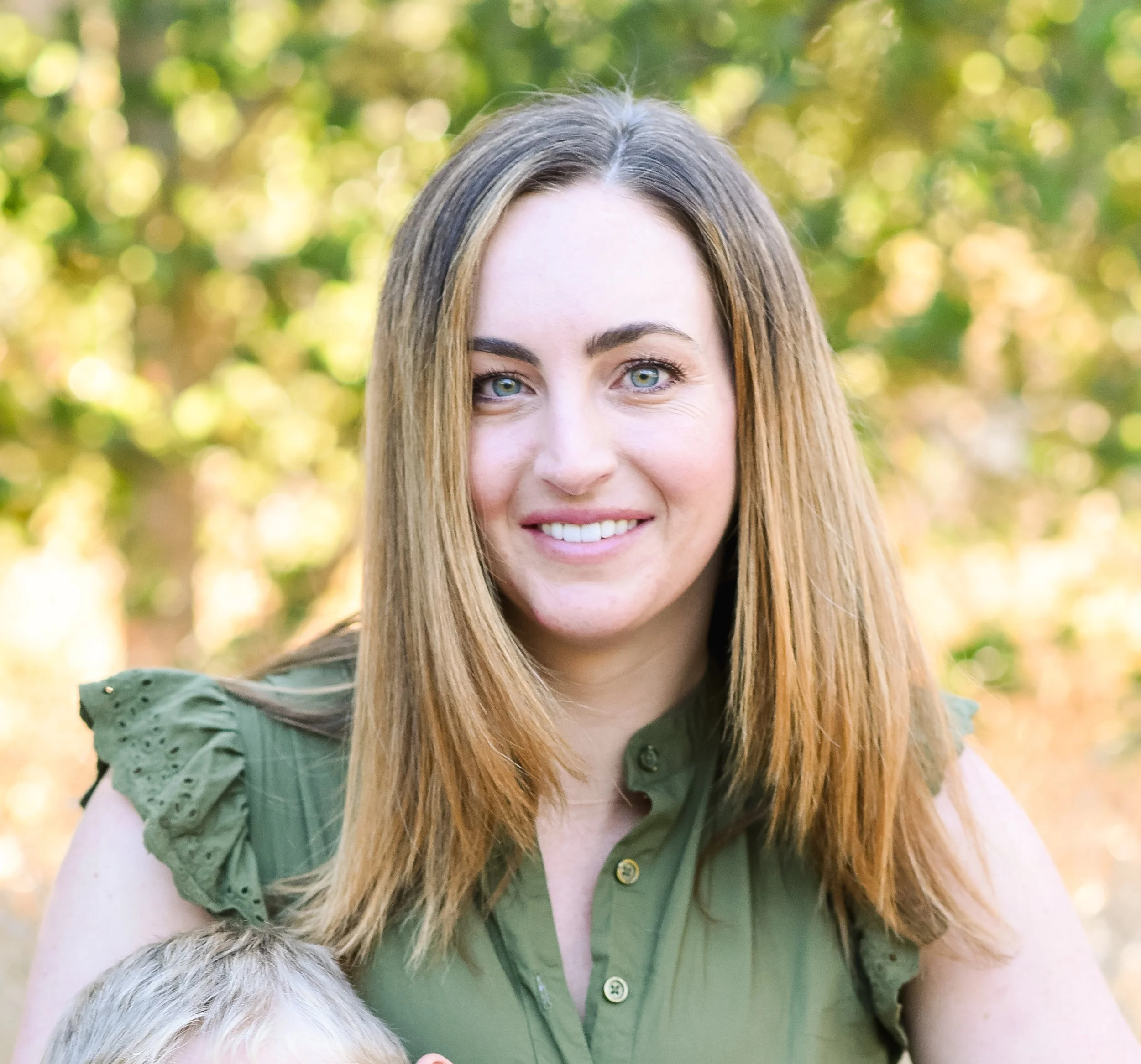 A woman with long, light brown hair and blue eyes smiling outdoors, wearing a green shirt with eyelet ruffles on the sleeves, with blurred green and yellow foliage in the background.