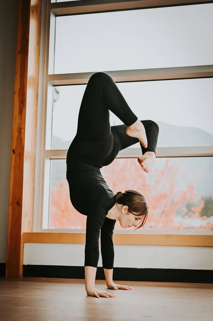 A woman practicing a headstand yoga pose indoors near large windows with a view of autumn trees.