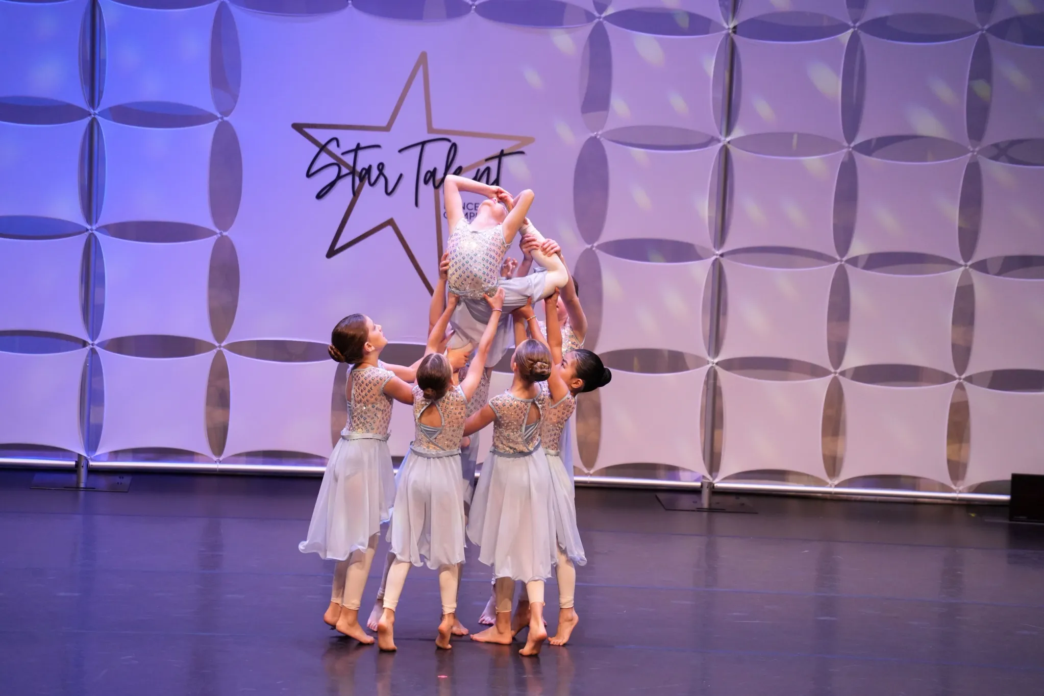 A group of young dancers dressed in beige costumes performing a ballet routine on stage, with some dancers holding one upside down and others supporting.